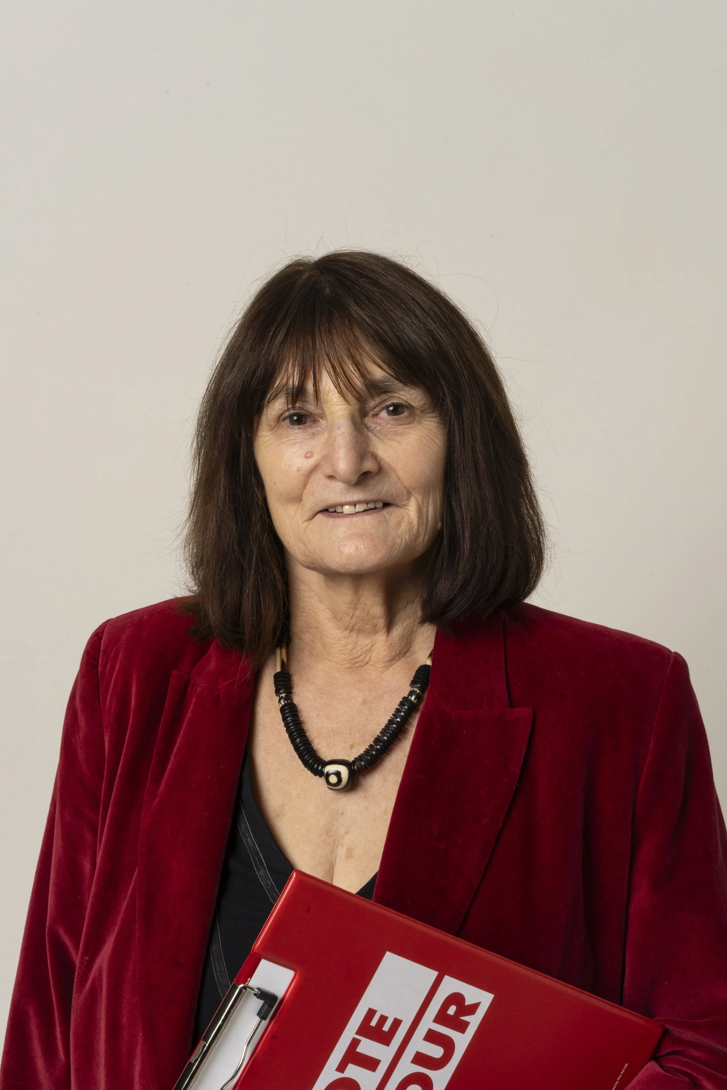 A woman with shoulder-length brown hair and bangs, wearing a red blazer, black top, and a black beaded necklace, holding a red folder.