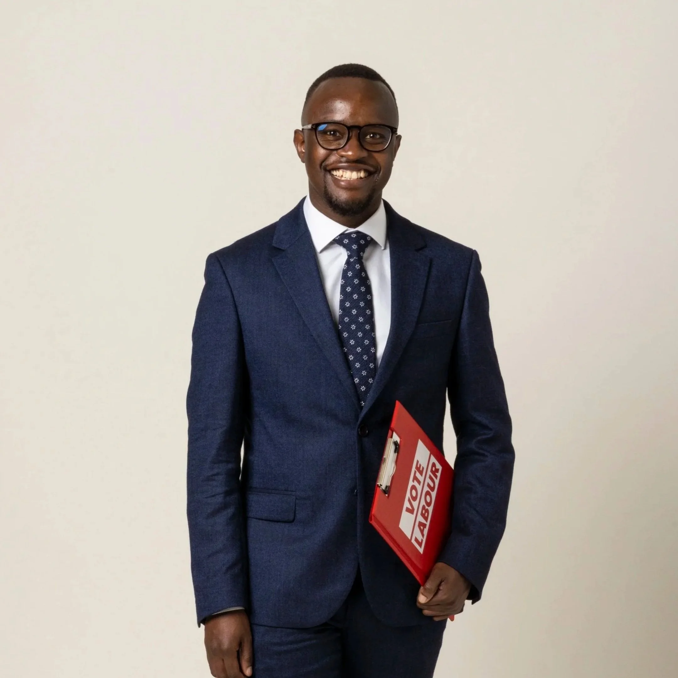 A smiling man wearing glasses, a dark suit, white shirt, and a pink patterned tie, standing against a plain light gray background.