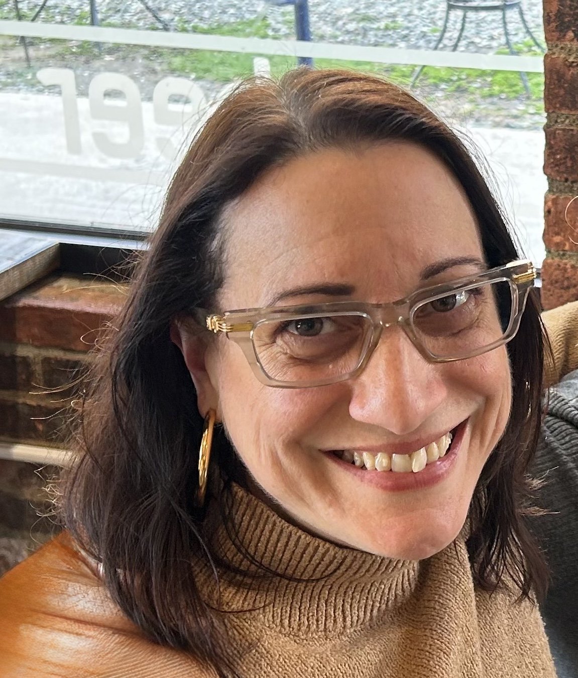 A man and woman hugging inside a bookstore, surrounded by wooden shelves filled with books. The man is wearing a baseball cap, glasses, and a white t-shirt. The woman is wearing glasses and a checkered shirt, smiling at the camera. The bookstore has warm lighting, green and brick walls, and framed pictures hanging on the walls.