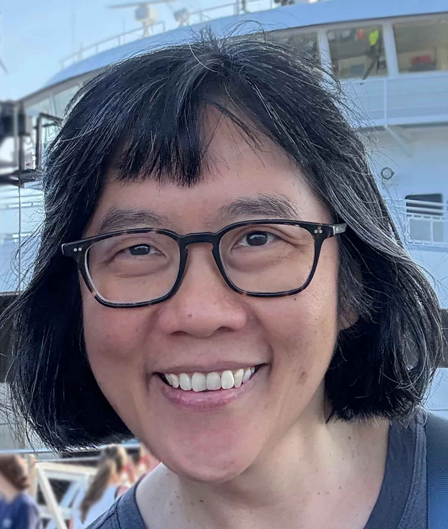 A woman with short dark hair and a nose piercing sitting on a couch, resting her head on her hand, in a room with natural light.