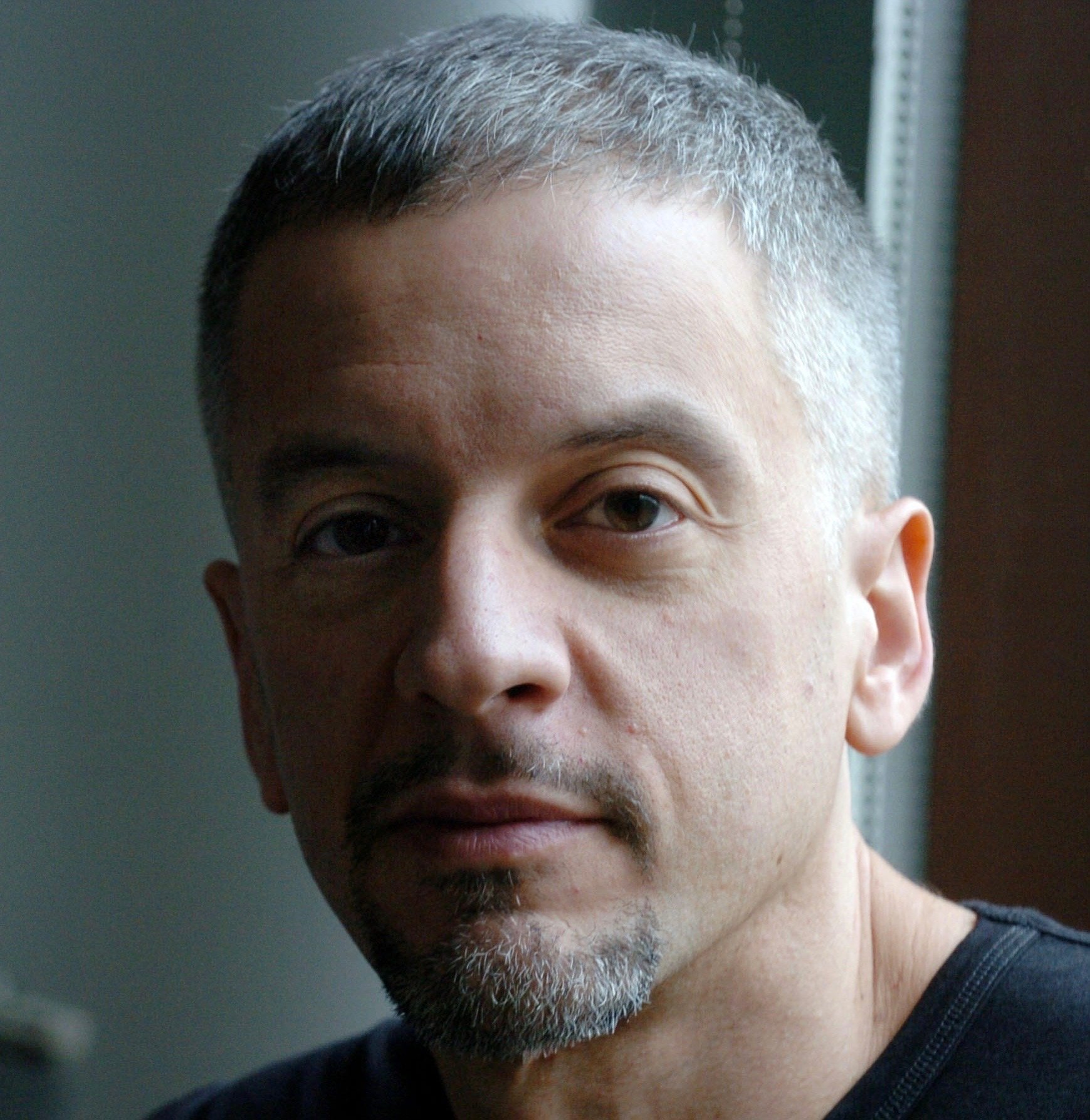 A woman with blonde hair and blue eyes smiling with her mouth open, holding a clear bottle with a white label and green text that reads 'Absinthe Prelude' and 'BiAncHe', standing inside a room with wooden walls and a coat rack in the background.