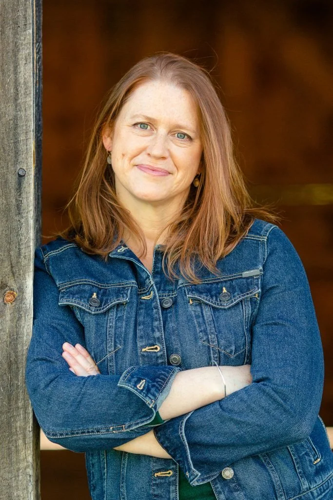 Close-up of an older woman with short gray hair, wearing blue glasses, a dark turtleneck, and a purple sweater, standing against a plain light-colored background.