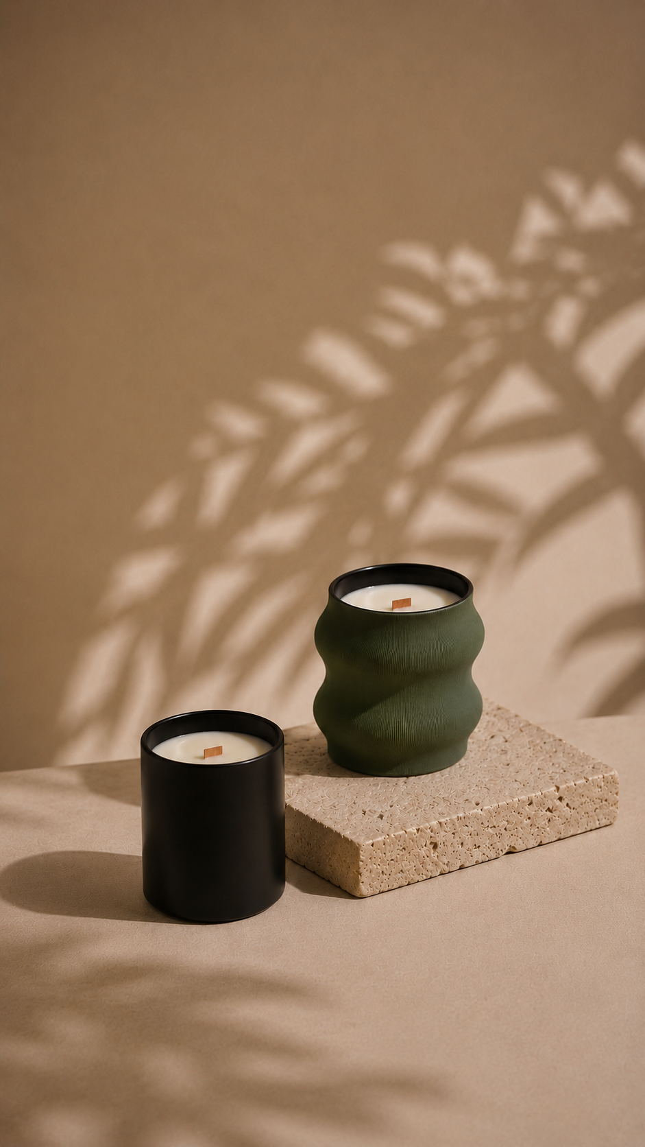 Two candles, one in a black container and one in a textured green container, placed on a beige surface with textured beige block, casting shadows of plant leaves on the wall behind.