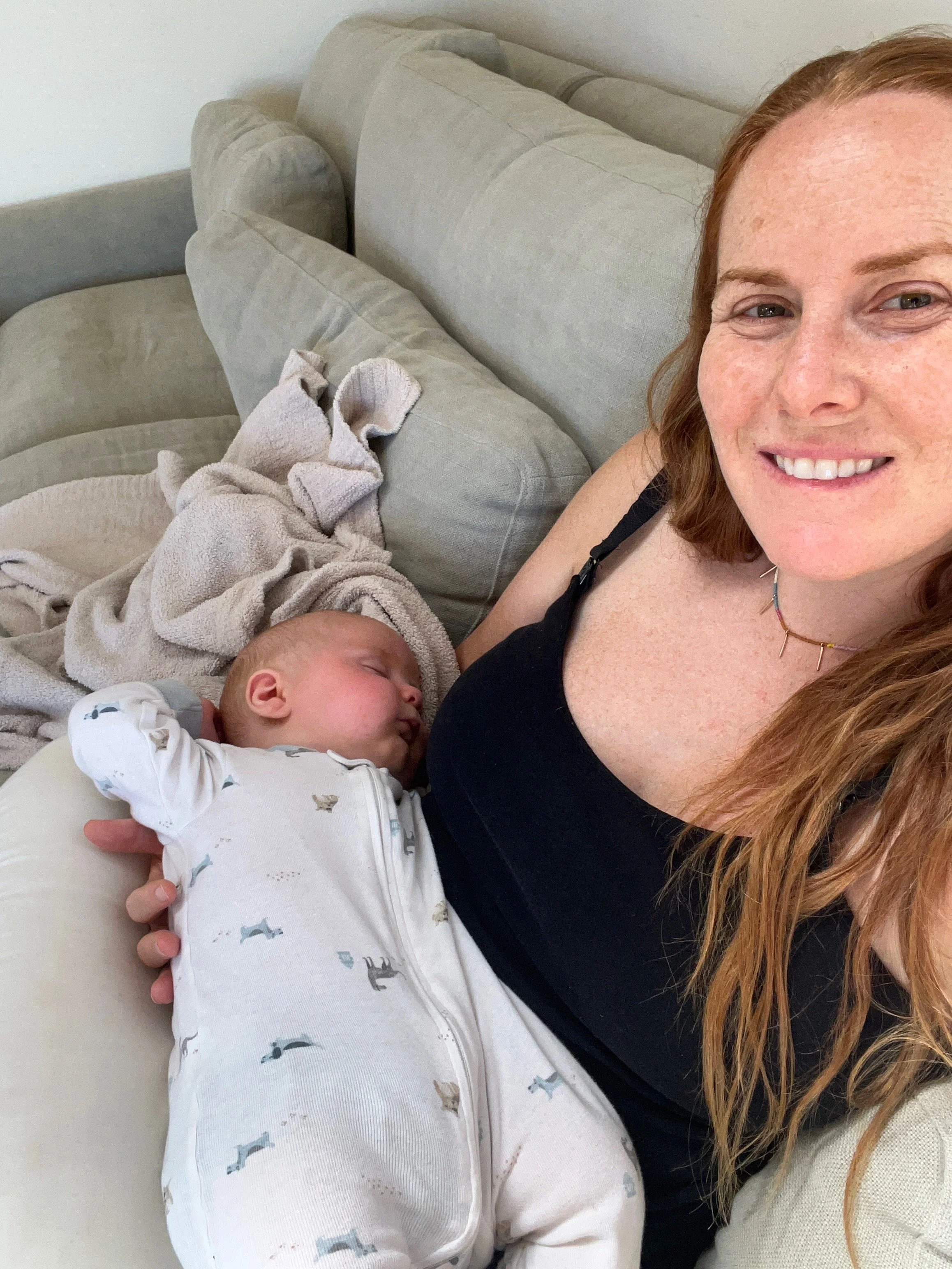 A woman with long red hair smiling and holding a sleeping baby in a white onesie with a dinosaur pattern, on a beige couch with cushions and a blanket.