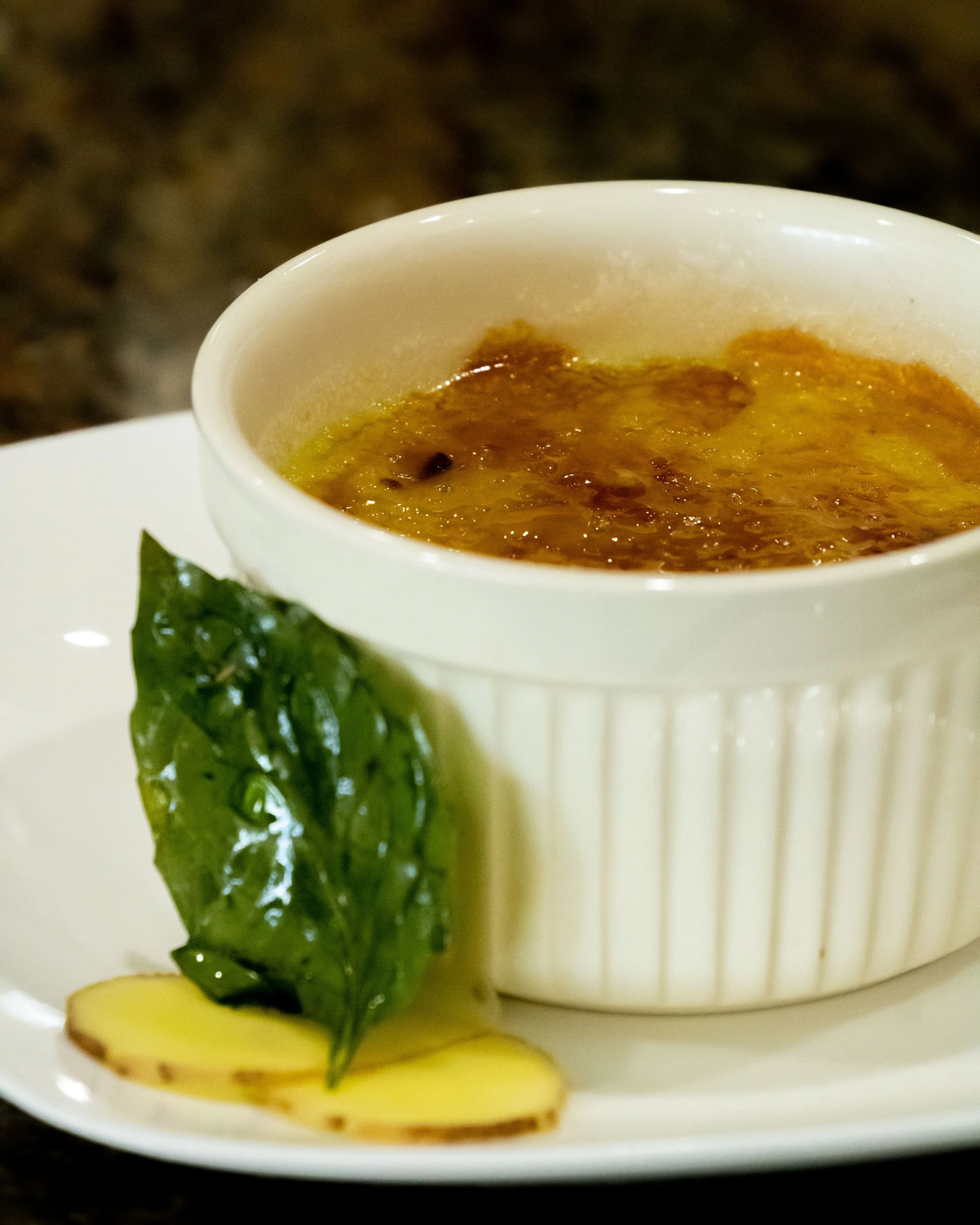 Close-up of a ramekin filled with baked crème brûlée, garnished with a green lettuce leaf and two slices of ginger on a white plate.