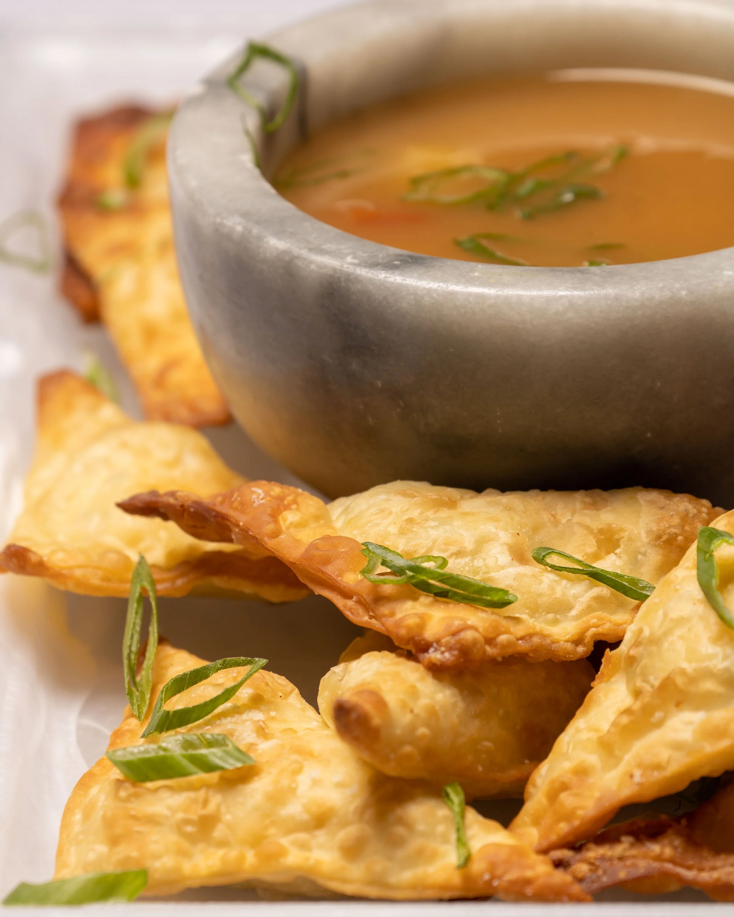 Close-up of fried wontons garnished with green onions, served with a bowl of orange dipping sauce containing chopped green herbs.