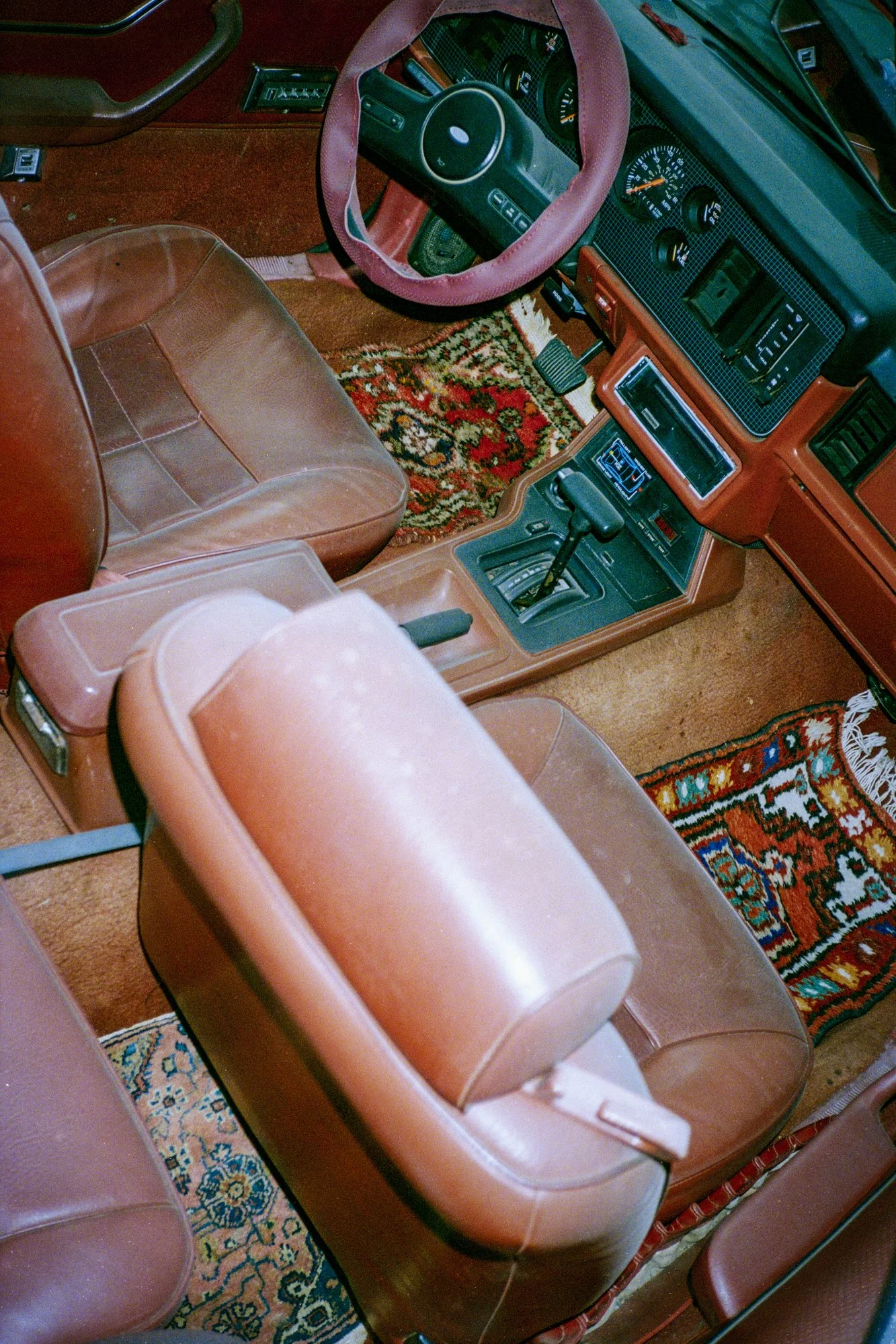 Interior view of a vintage car showing brown leather seats, a dashboard with various gauges, a gear shift, a pink steering wheel cover, and colorful patterned rugs on the floor.