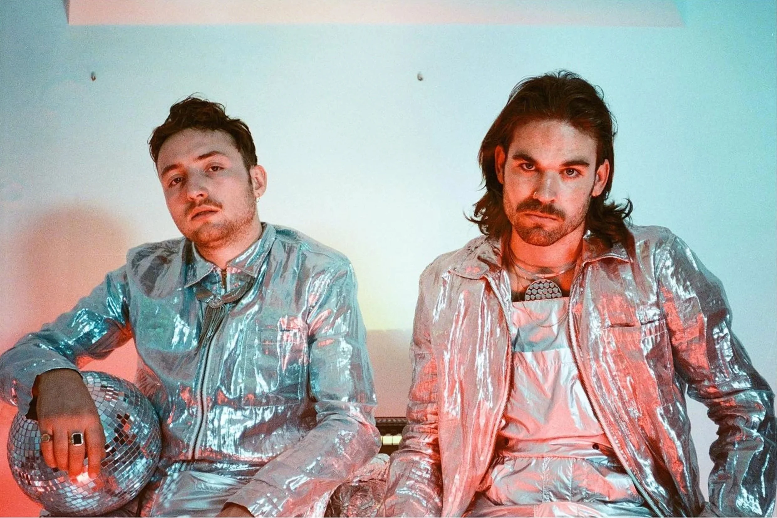Two men sitting against a plain white wall, wearing shiny metallic jackets, one holding a disco ball, with a colorful lighting effect.