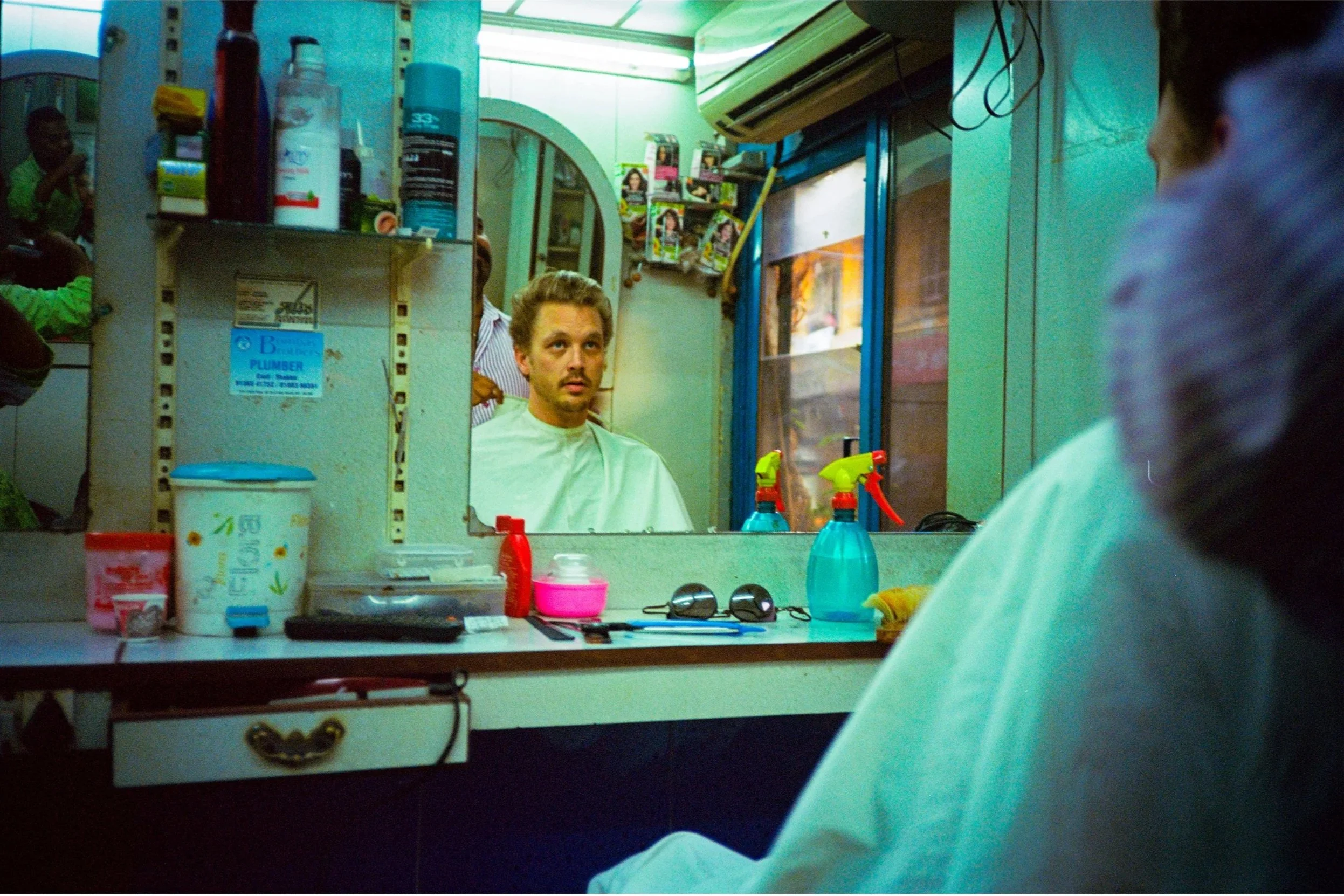 A man getting a haircut in a barbershop, looking at his reflection in the mirror, with barber tools and spray bottles on the counter.