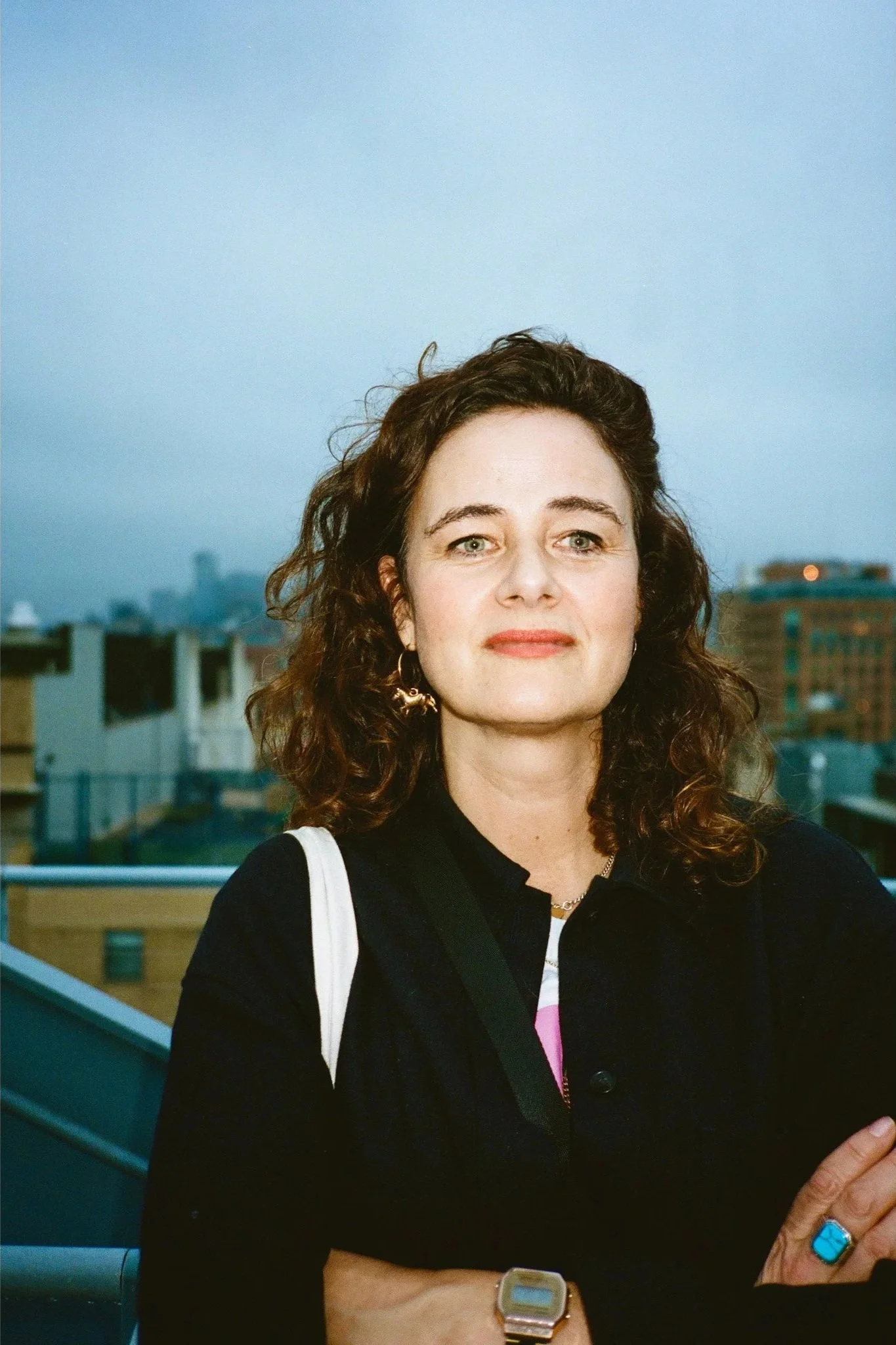 A woman with curly brown hair and fair skin standing outdoors on a city rooftop at dusk, looking at the camera, with city buildings and a cloudy sky in the background.