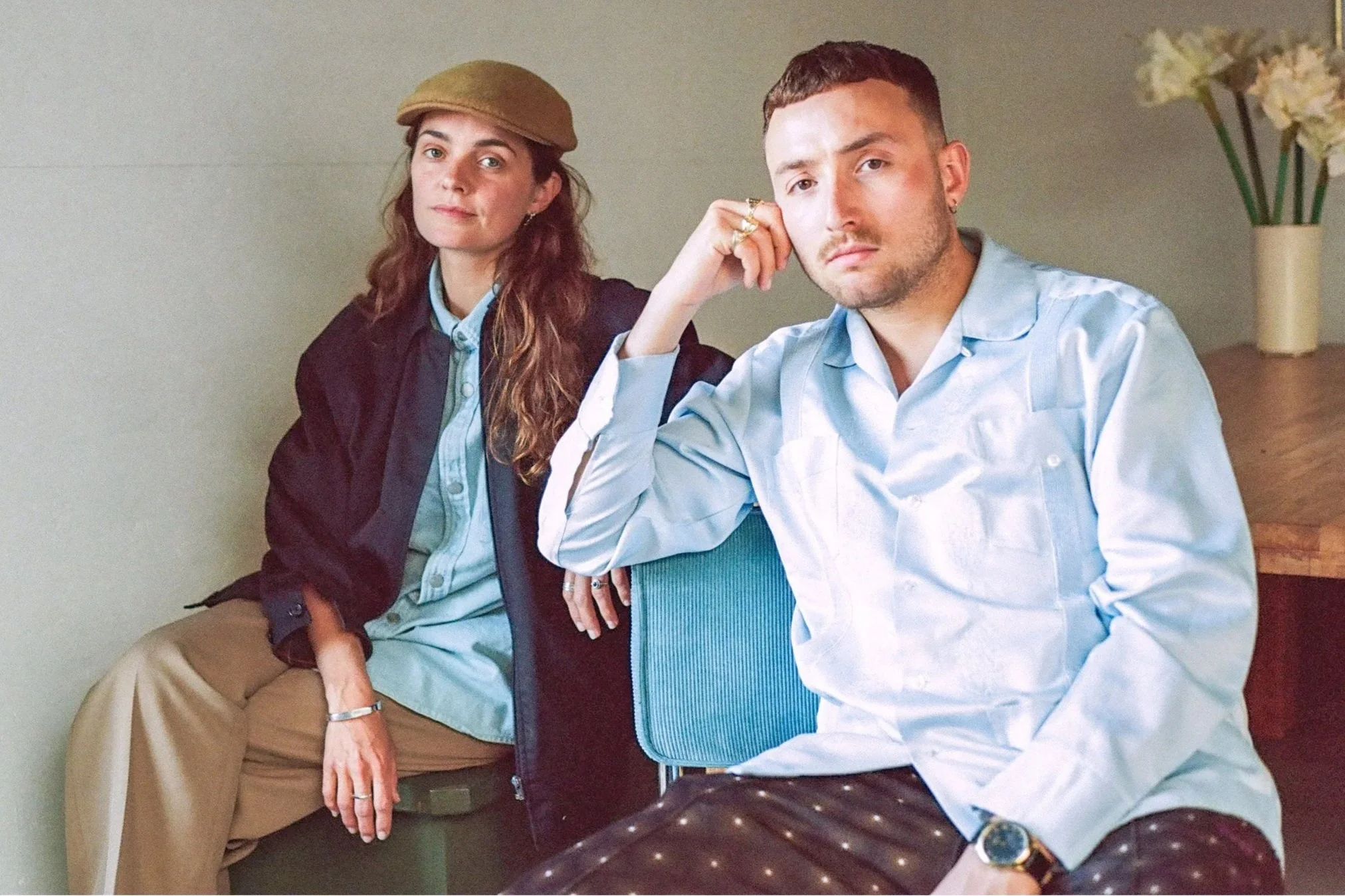 A young man and woman sit in a room, looking at the camera. The woman has long, wavy hair, wearing a beige hat, a burgundy jacket, and beige pants. The man has short hair, wearing a light blue shirt, patterned pants, and a watch.