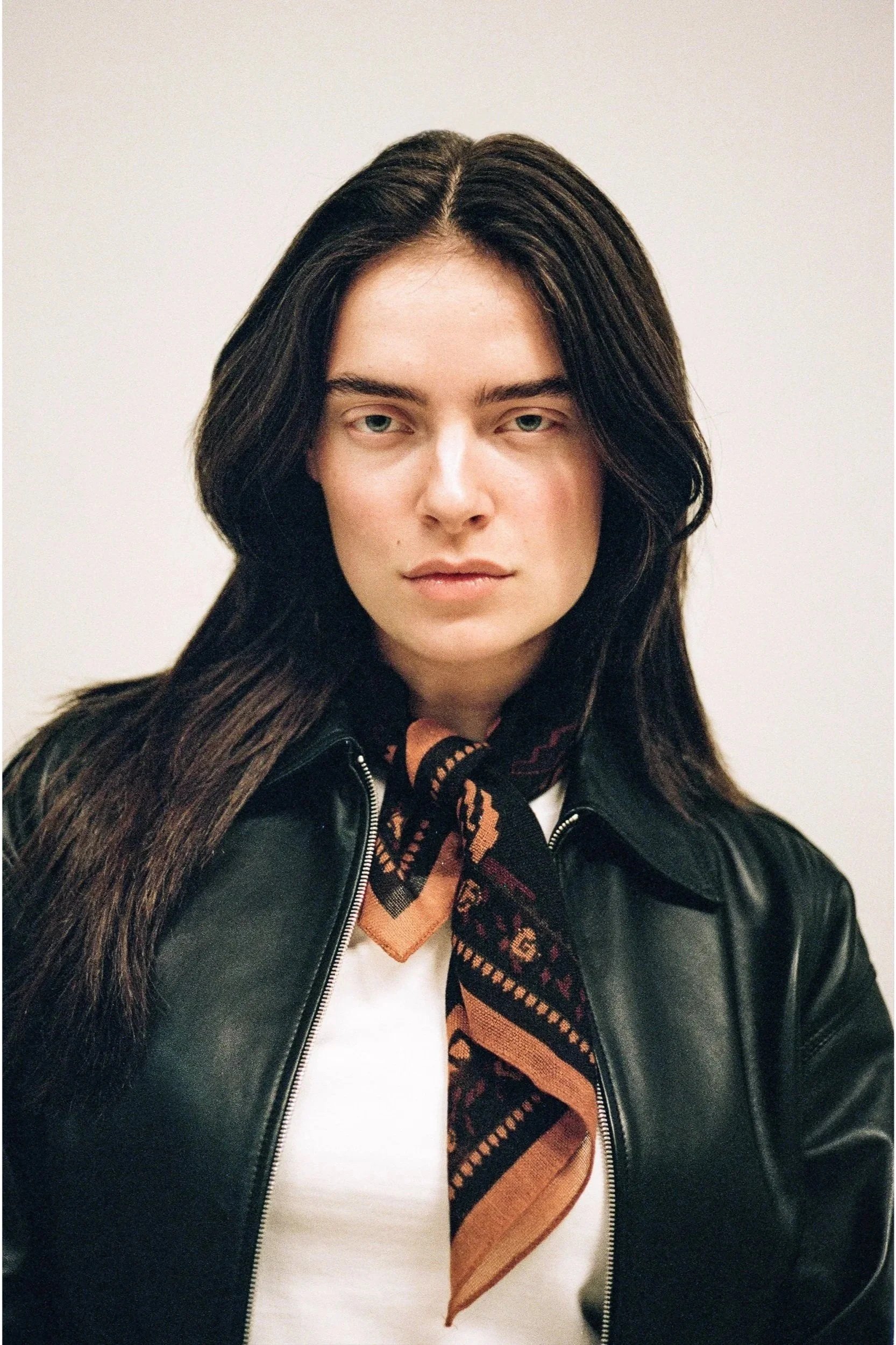 A woman with long dark hair, wearing a black leather jacket and a patterned scarf, looking at the camera against a plain white background.