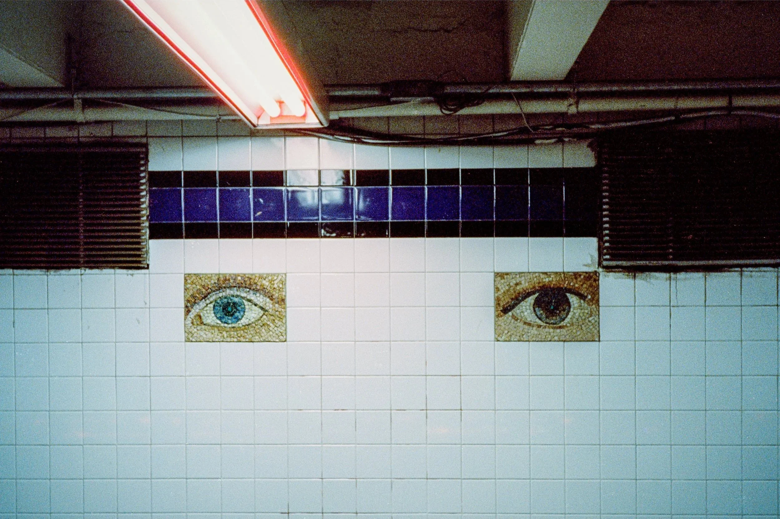 A tiled wall with two eyes mosaics, above a black horizontal stripe, with a red neon sign on the top left corner.