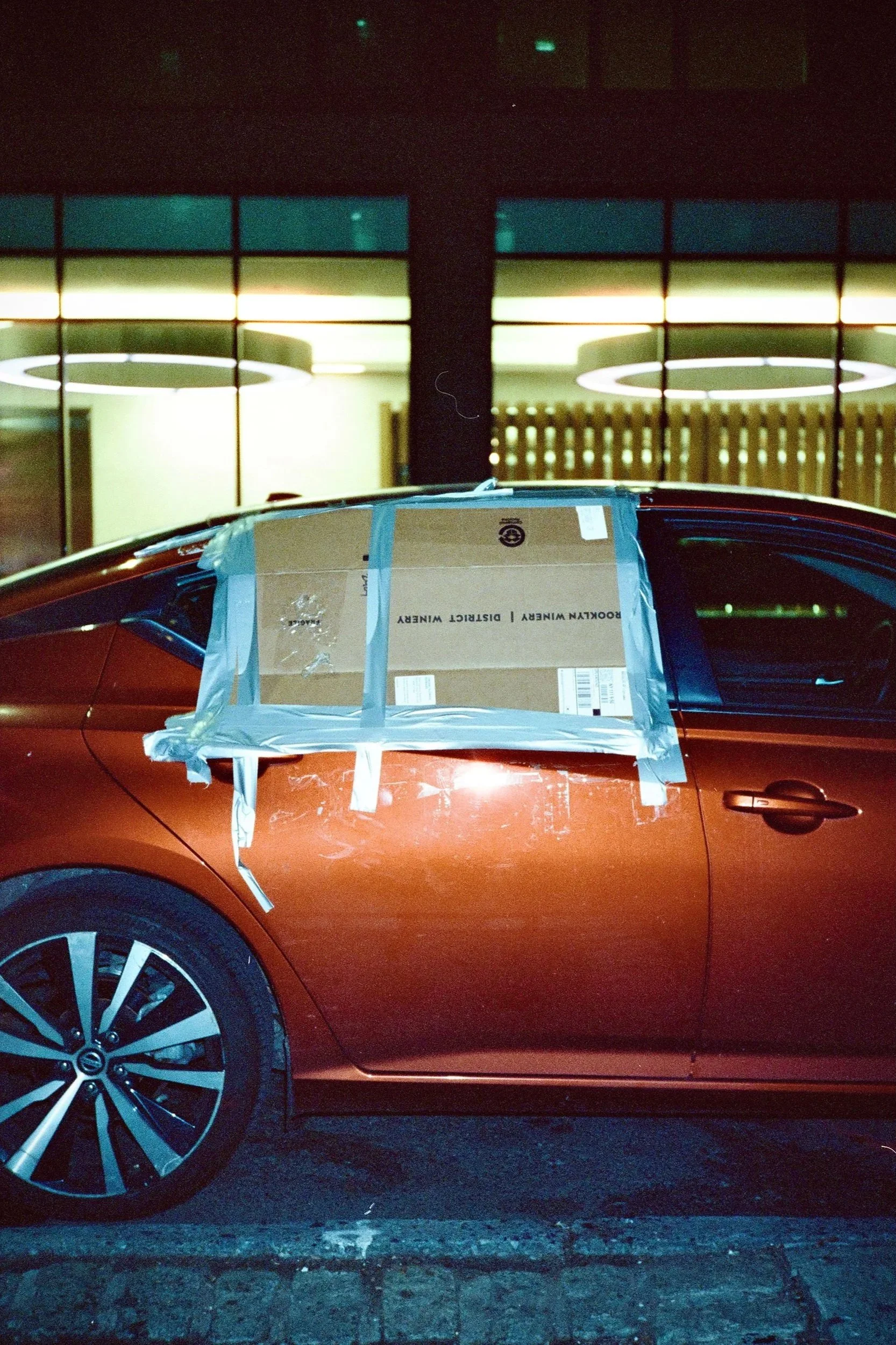 A red car with a cardboard box taped on the driver's side window, covered with blue painter's tape, parked outside at night, with illuminated indoor lights and large round ceiling fixtures visible in the background.