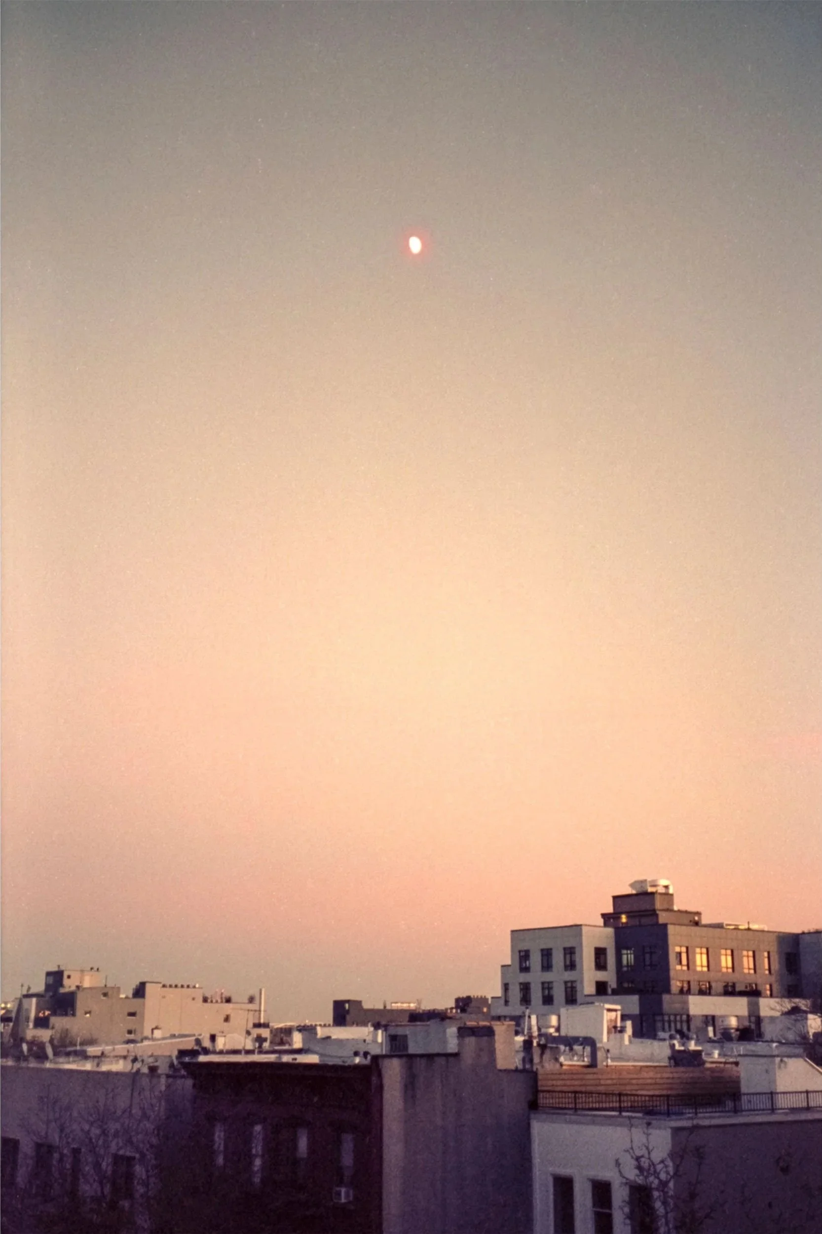 Sky at dusk showing a bright celestial object, possibly the moon or a planet, above city rooftops with buildings and windows reflecting the sunset.