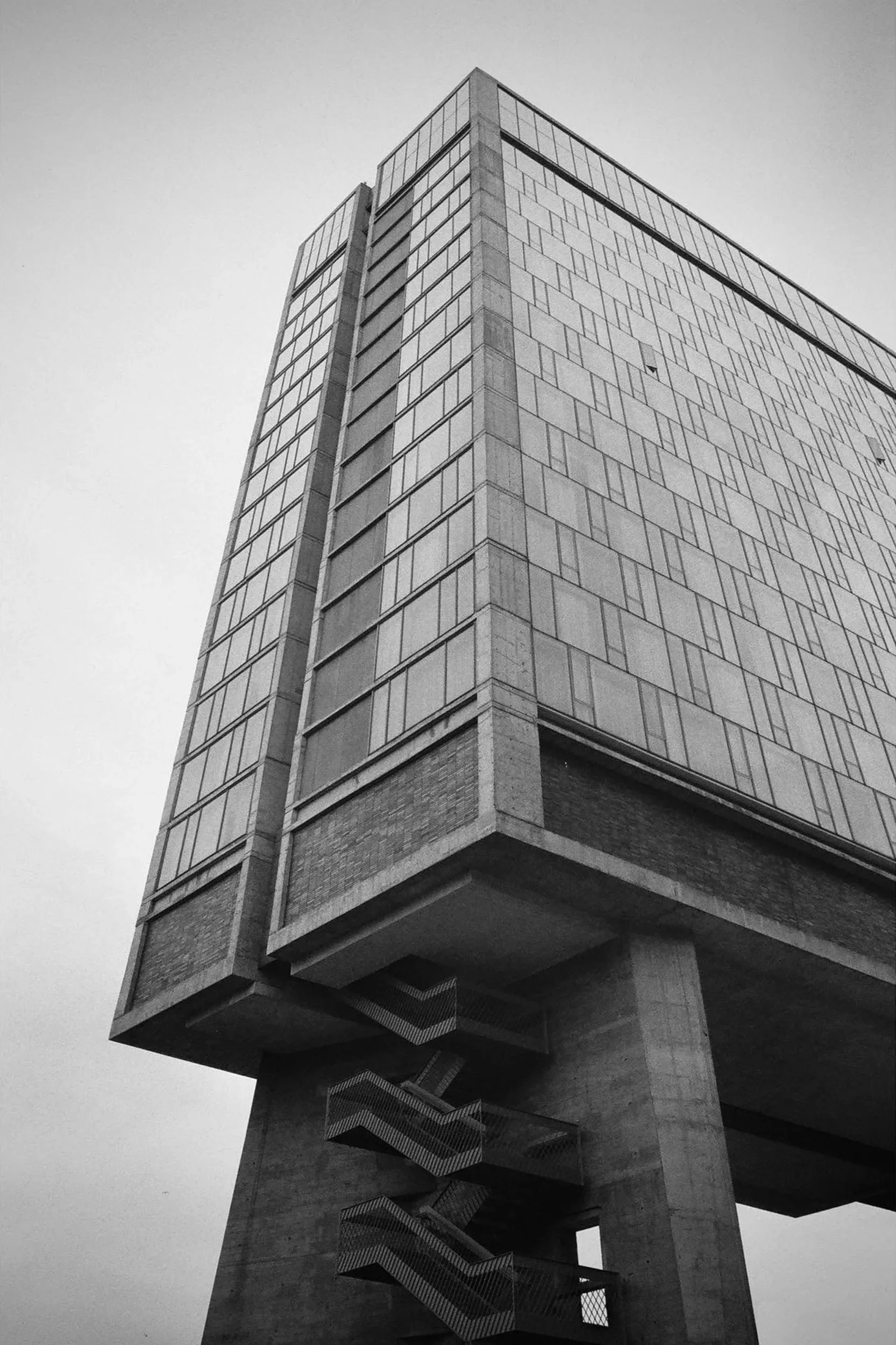 Black and white photo of a modern tall office building with glass windows, viewed from below, with a staircase visible underneath.