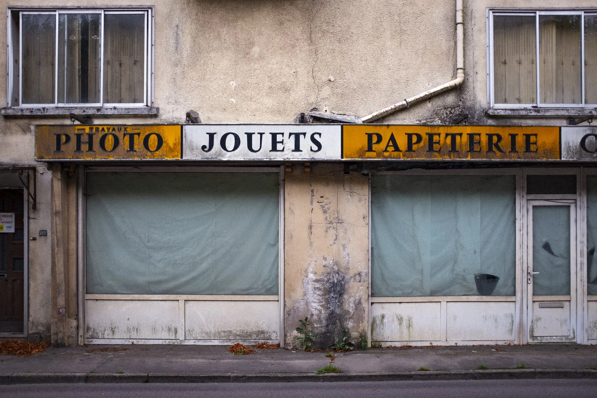 An old storefront with yellow and white sign that says 'Photo Jouets Papeterie' showing peeling paint and dirt, closed windows covered with white paper, and a dirty sidewalk with fallen leaves.