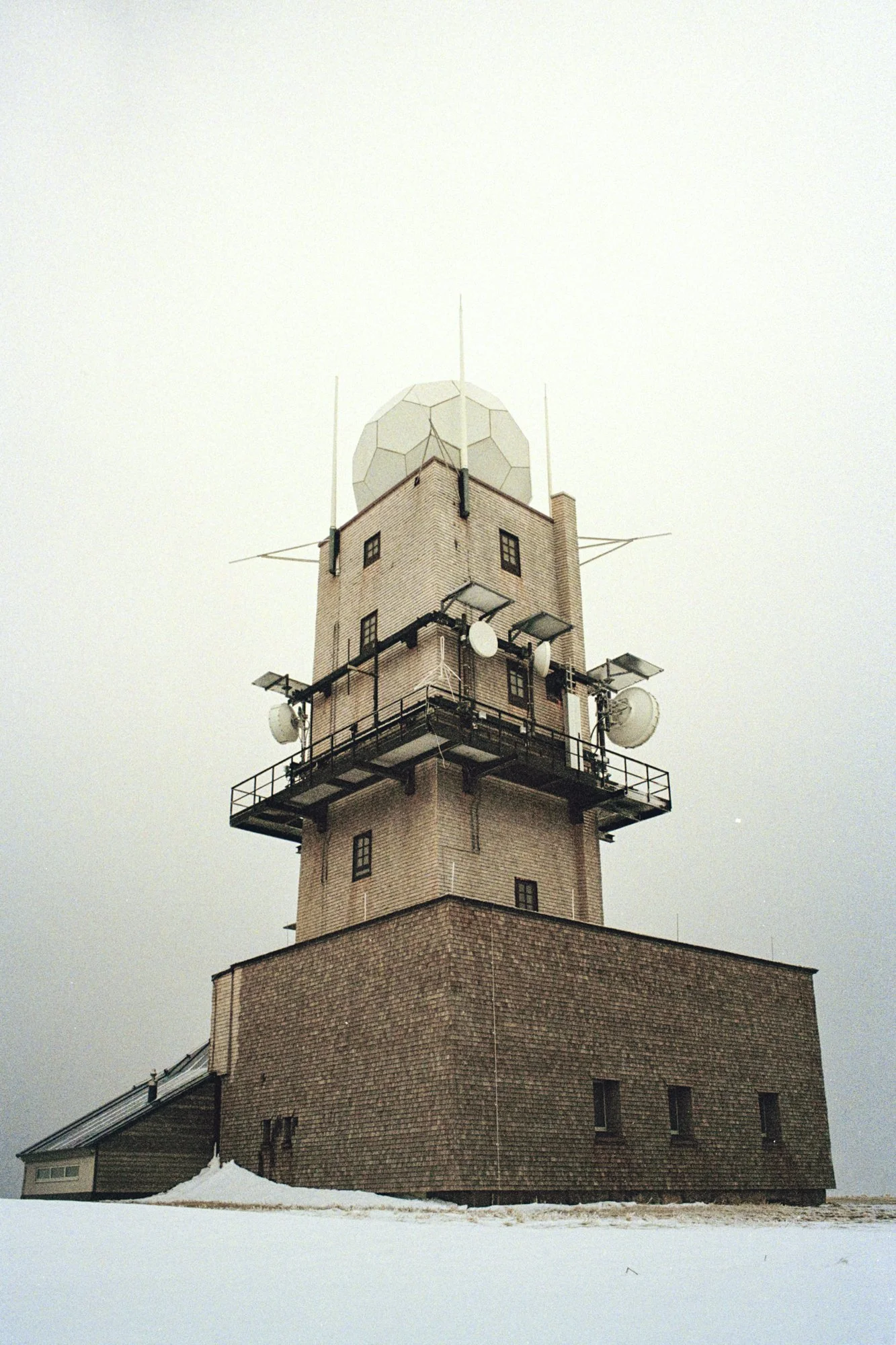 A tall brick tower with multiple communication antennas and satellite dishes on the upper levels, standing on snow-covered ground under an overcast sky.