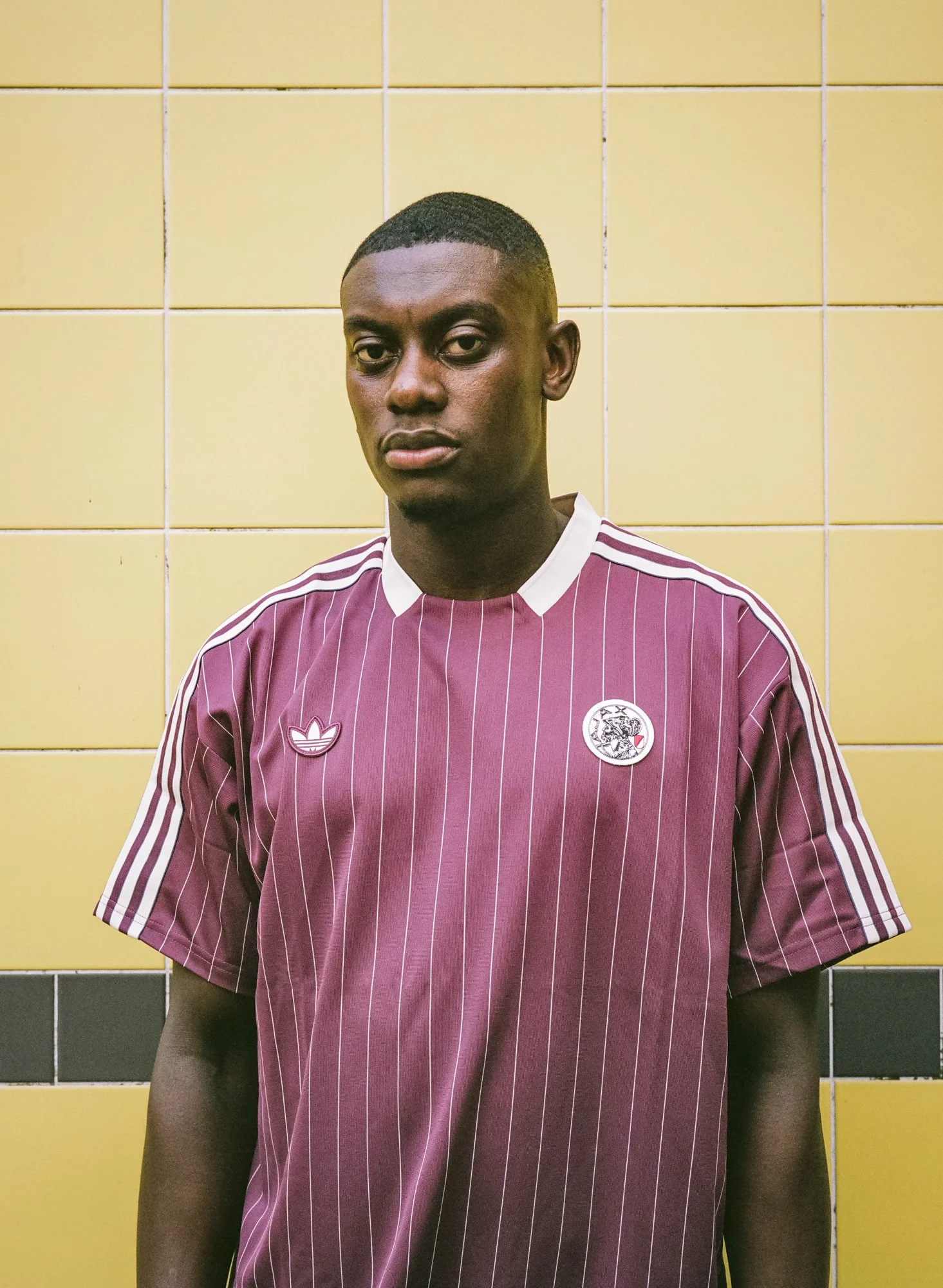 A young man with dark skin, short hair, and a serious expression standing against a yellow tiled wall. He is wearing a maroon sports jersey with white pinstripes, featuring a white Adidas logo and a circular emblem.