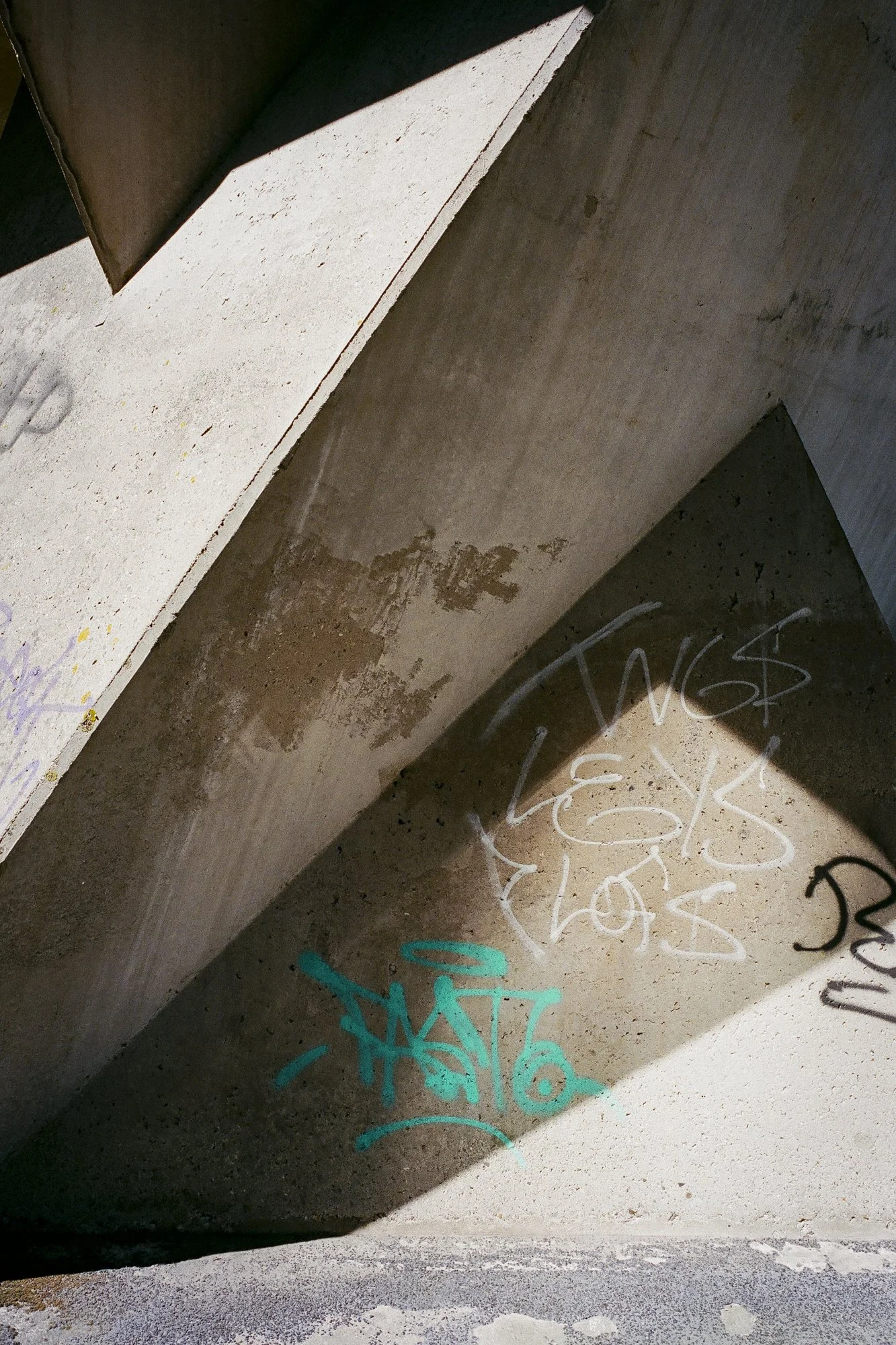Graffiti tags in white and teal spray paint on a concrete wall, with a shadow cast over part of the wall.