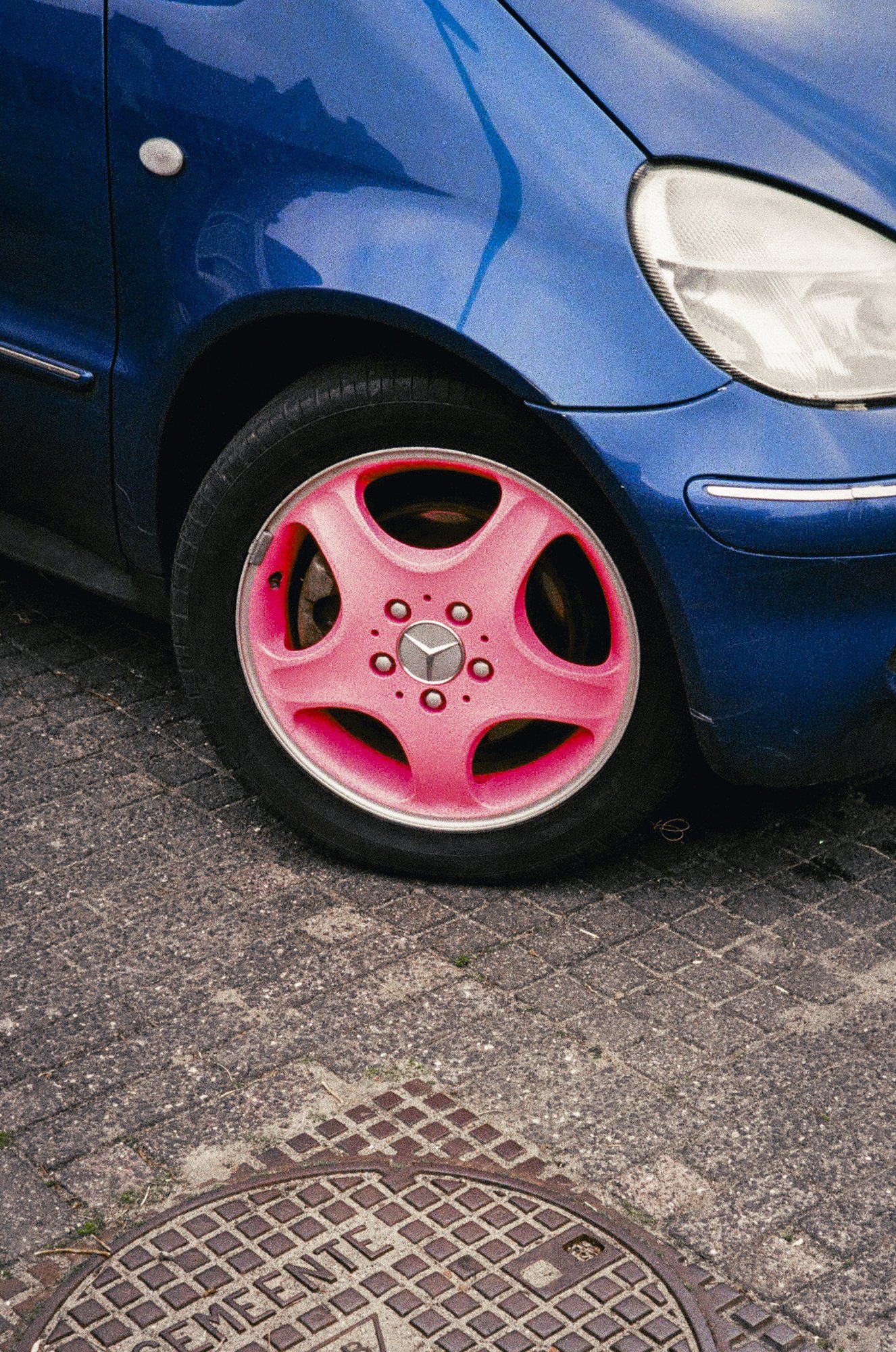 Close-up of a blue car with a pink Mercedes-Benz wheel rim parked on a street with a manhole cover in the foreground.