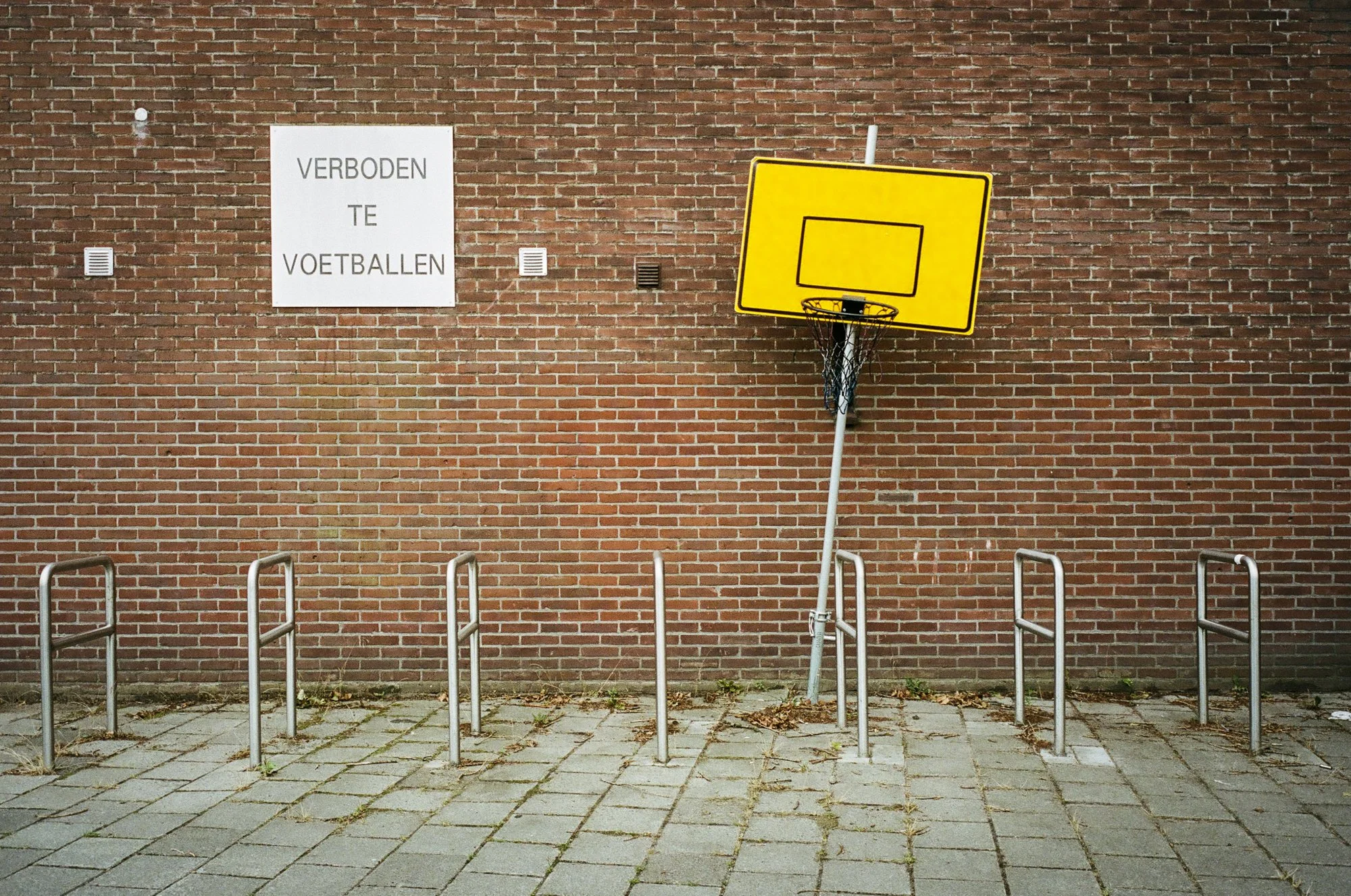 Empty outdoor basketball court with a yellow backboard and hoop, metal bike racks, and a sign on a brick wall that says 'Verboden te voetballen' (No soccer allowed in Dutch).