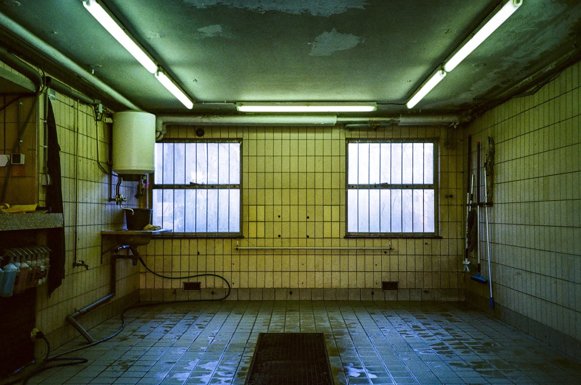 An empty room with two windows, tiled walls, fluorescent lights, a sink, cleaning tools, and a floor drain.