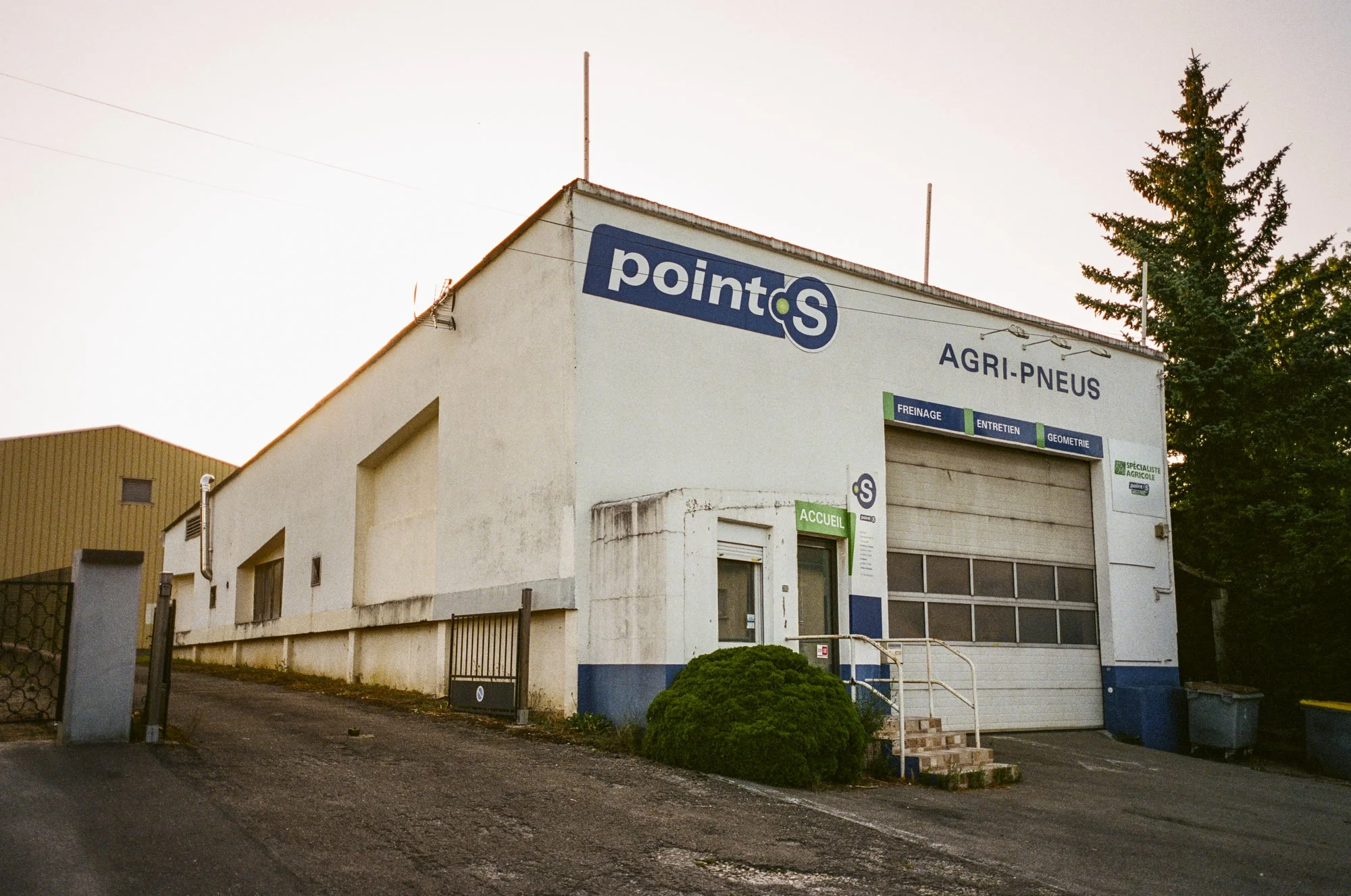 A white building with blue and green signs, including 'point S' and 'AGRI-PNEUS', featuring service signs for brake, maintenance, and geometry work, with a small entrance labeled 'ACCUEIL' and a garage door, surrounded by trees and a partly cloudy sk