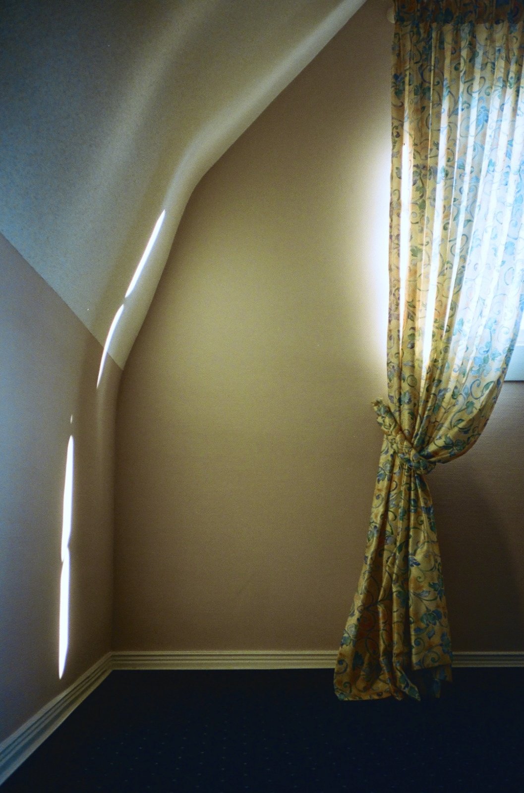 A corner of a room with a sloped ceiling, beige walls, a dark carpet, and a window with light-colored, floral-patterned curtains tied back, allowing light to enter.