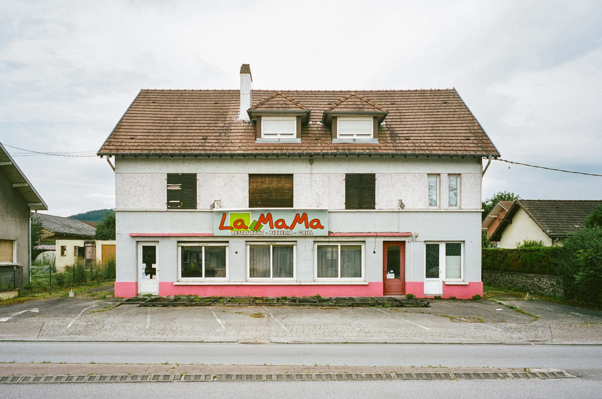 A two-story building with a restaurant named La MaMa on the ground floor, featuring a painted sign, windows, and a door. The building has a brown tiled roof and white walls with pink accents at the bottom.