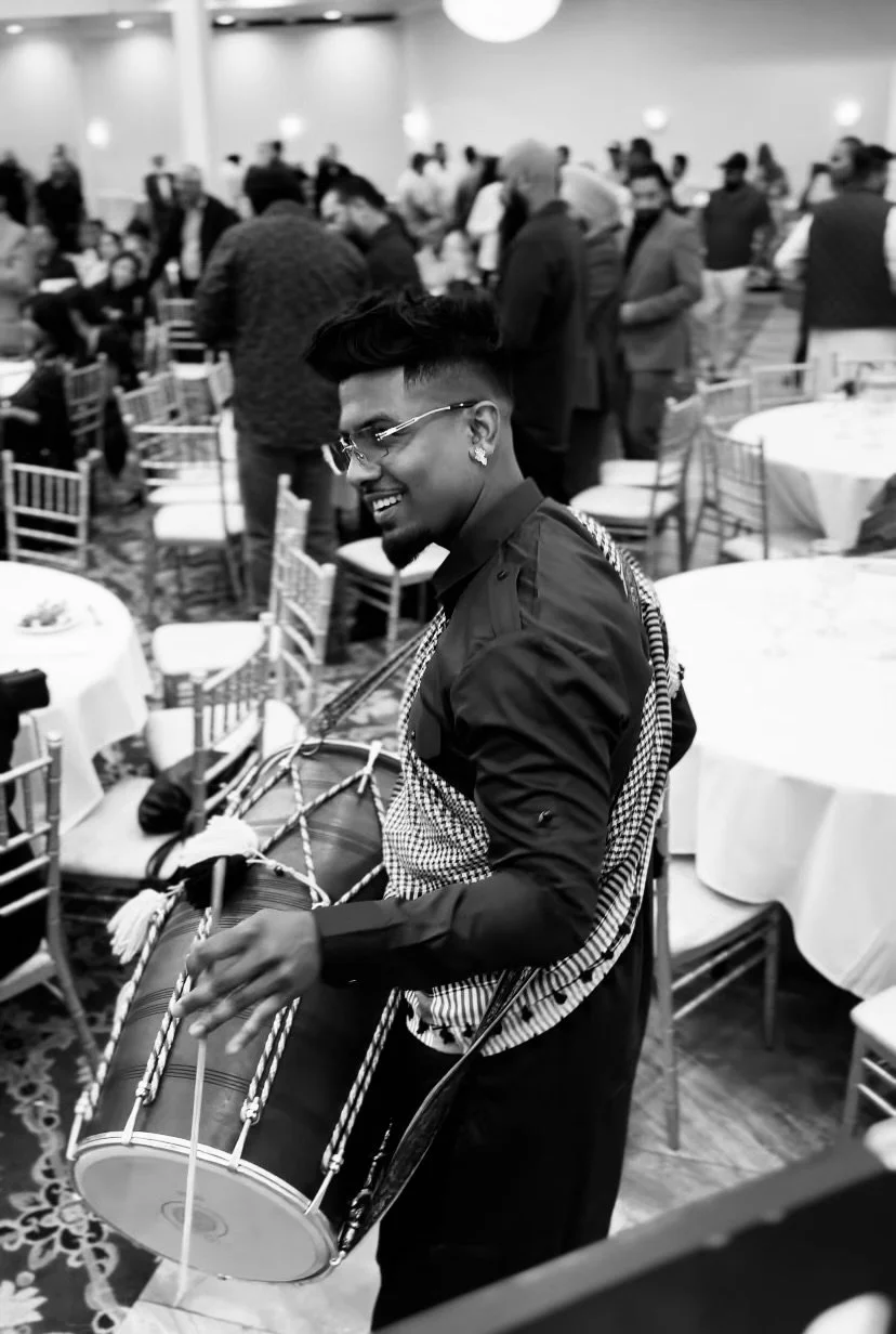 Smiling man playing a dhol drum at an event with people seated at tables in the background.