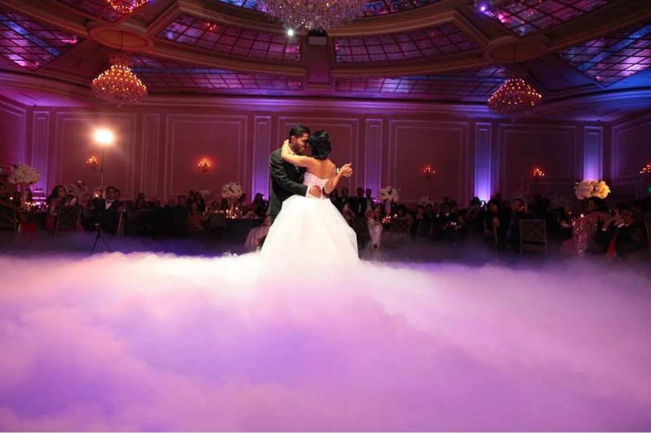 Bride and groom dancing on a cloud-filled floor in an elegant ballroom with chandeliers and purple lighting.