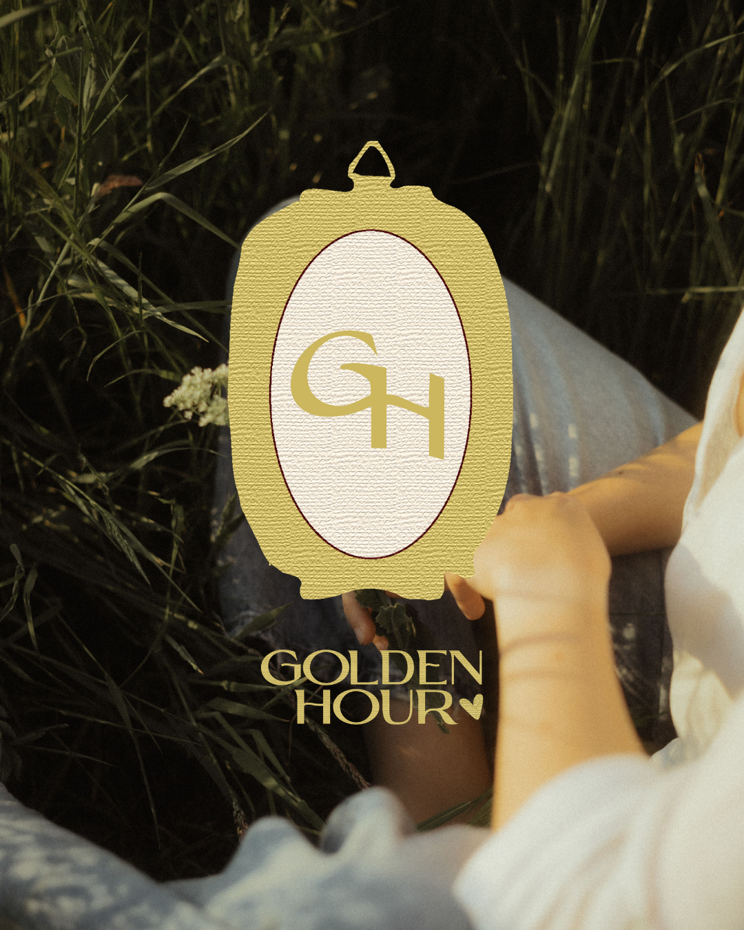 A person sitting outdoors in a grassy area holding a sign with the logo ‘GH’ and the words ‘Golden Hour’ below it.