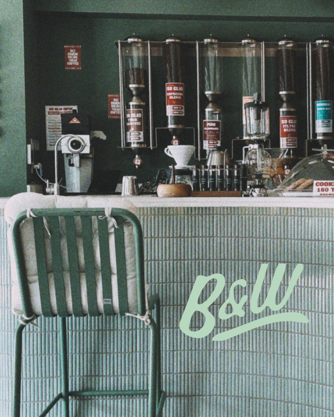 Coffee shop counter with bins of coffee beans, a vintage coffee grinder, a metal coffee dripper, and a glass display case of baked goods. A green chair is in front of the counter, and the wall behind is green with signs.