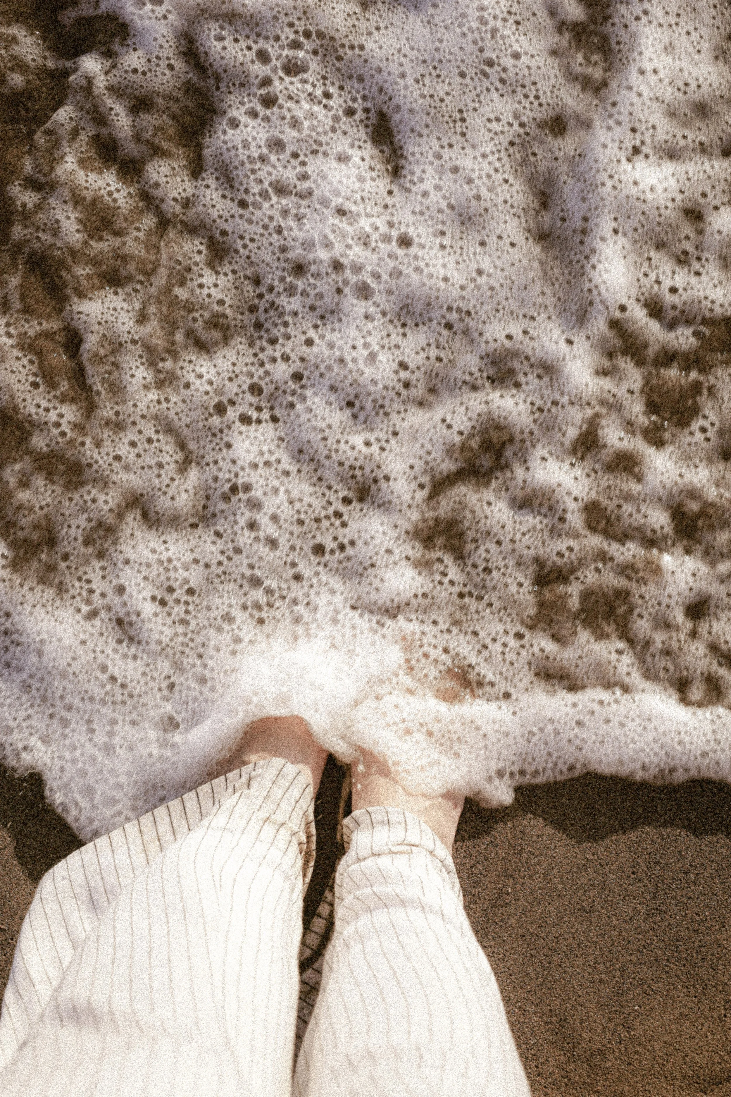 A person wearing cream-colored, striped pajama pants standing on a sandy beach with foamy sea waves.