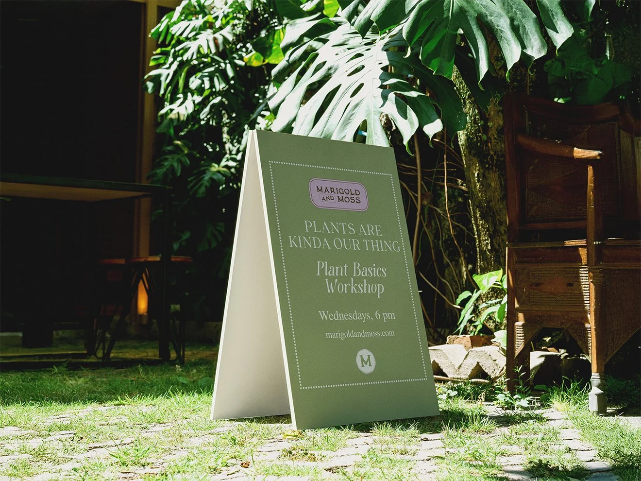 Outdoor signboard promoting a plant basics workshop by Marigold and Moss, held on Wednesdays at 6 pm, in a garden area with large tropical leaves and wooden furniture.