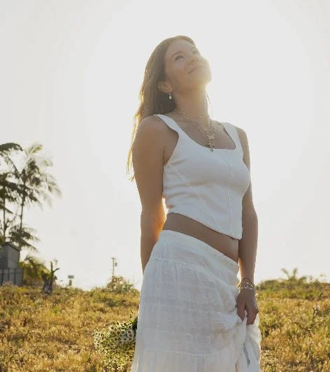 Woman in white outfit standing outdoors at sunset, smiling and looking up.
