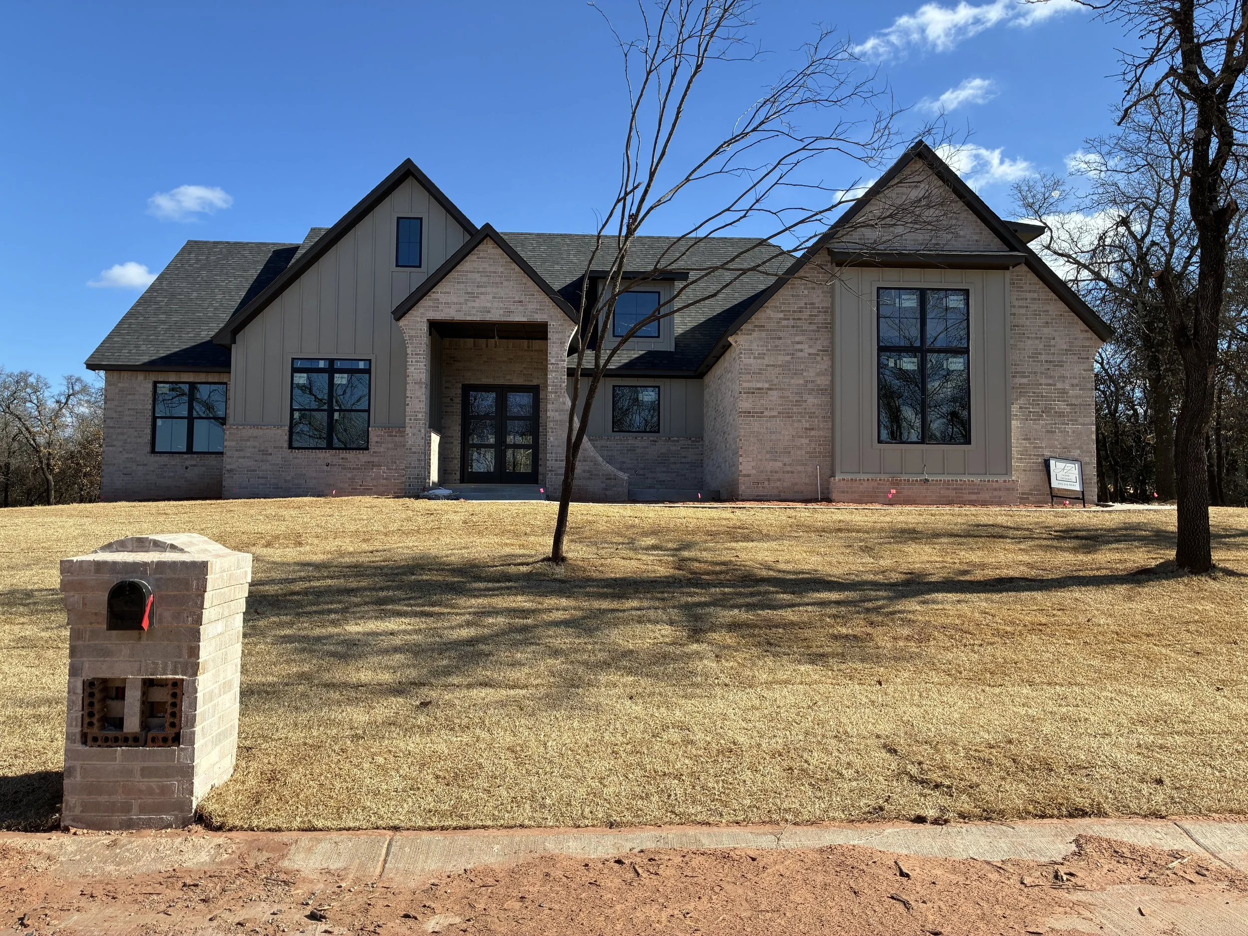 A modern brick house with large black-framed windows, a gray shingled roof, and a small porch in a yard with a grass lawn and leafless trees. A brick mailbox is in the foreground, and a yard sign is visible near the house.