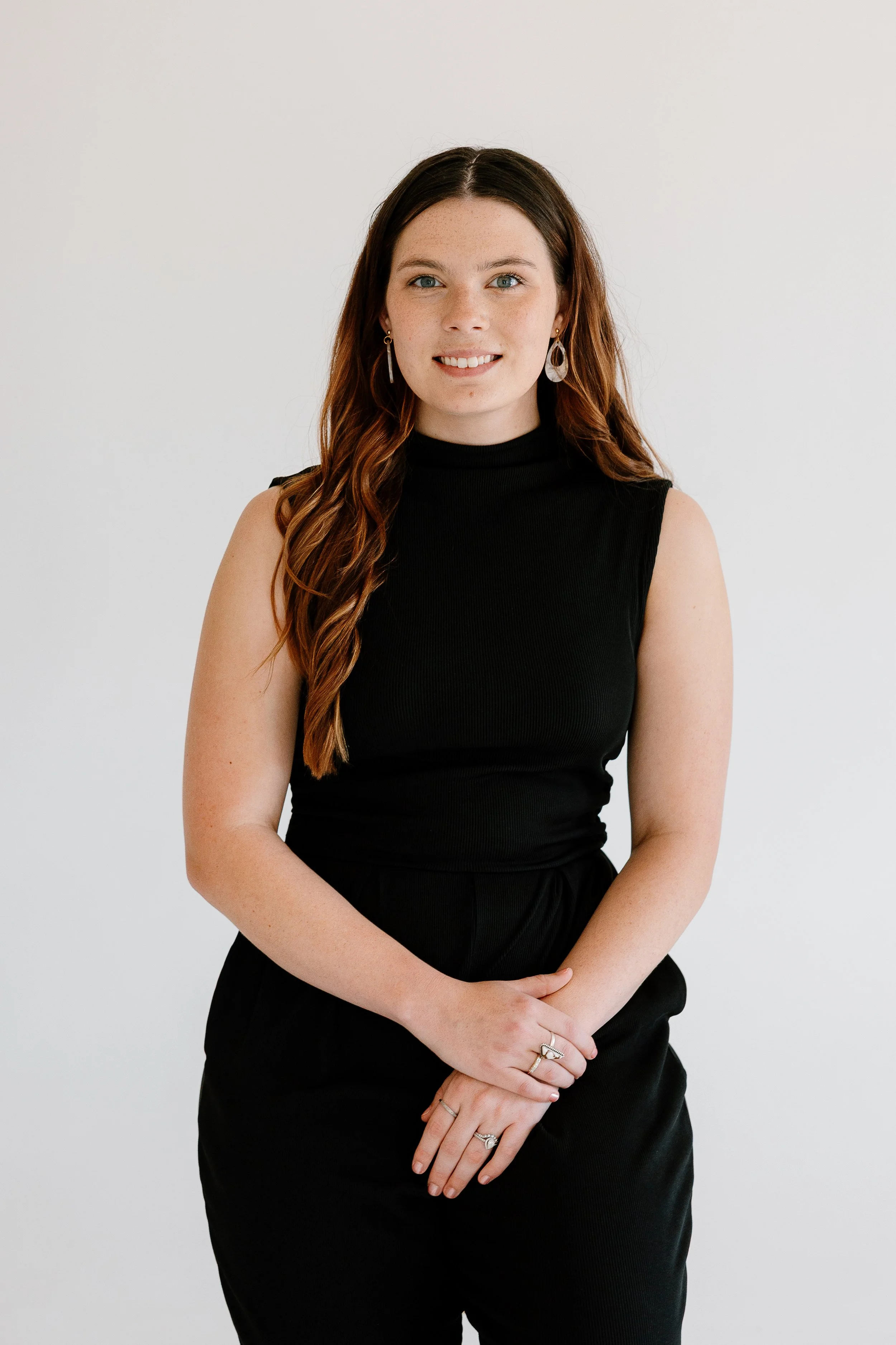 Portrait of a young woman with long wavy brown hair, wearing a sleeveless black top, and multiple rings and earrings, standing against a plain white background, smiling softly.