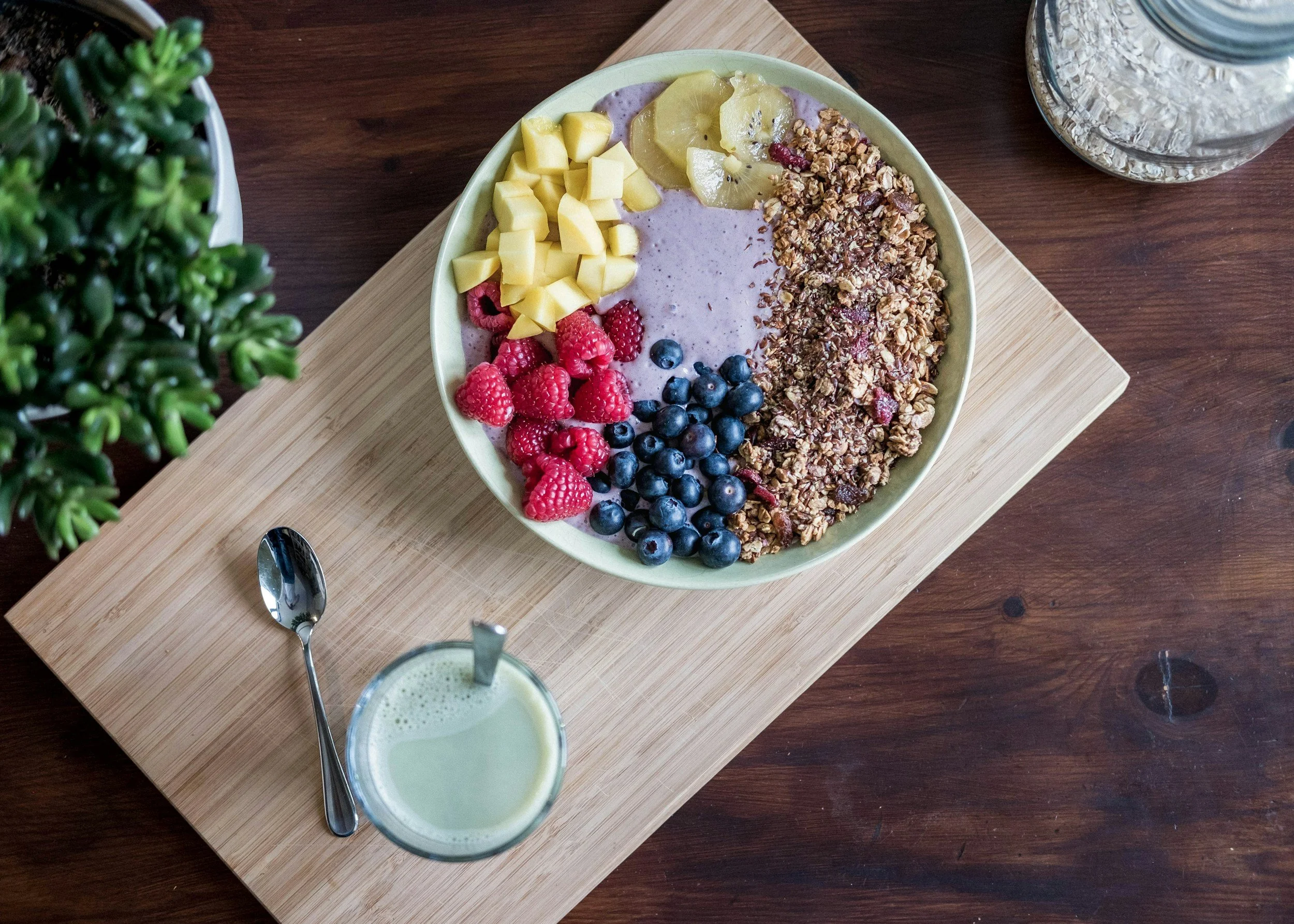 Yogurt bowl with granola, berries, and pineapple, and a side of juice, all sitting on top of a wooden cutting board.