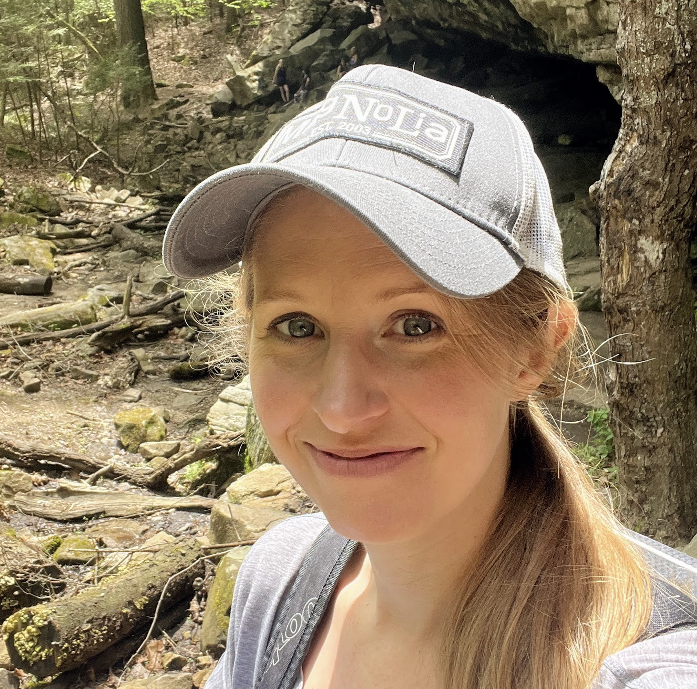 A woman with light skin, brown hair, and blue eyes smiling while wearing a gray baseball cap and a gray shirt, in a forested area with rocks, trees, and a small waterfall in the background.