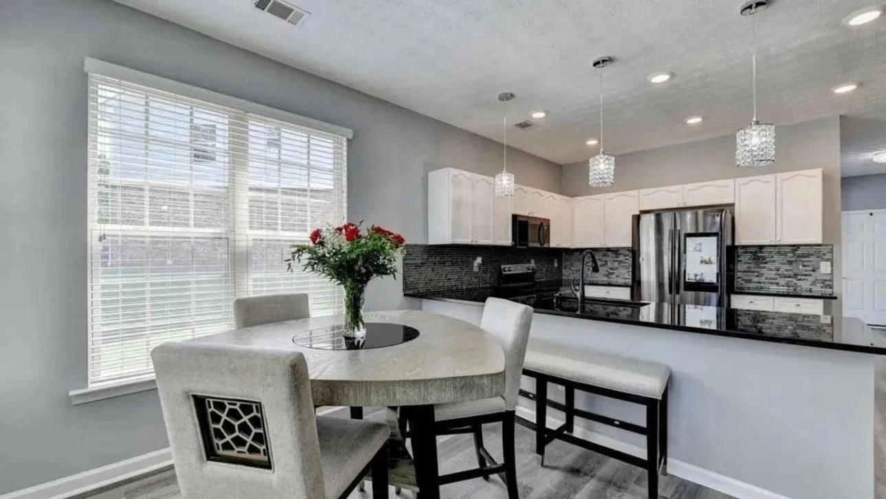 Modern kitchen with white cabinets, black countertops, and stainless steel appliances. A round dining table with chairs and a vase of red flowers in the foreground. Large window with blinds allows natural light.