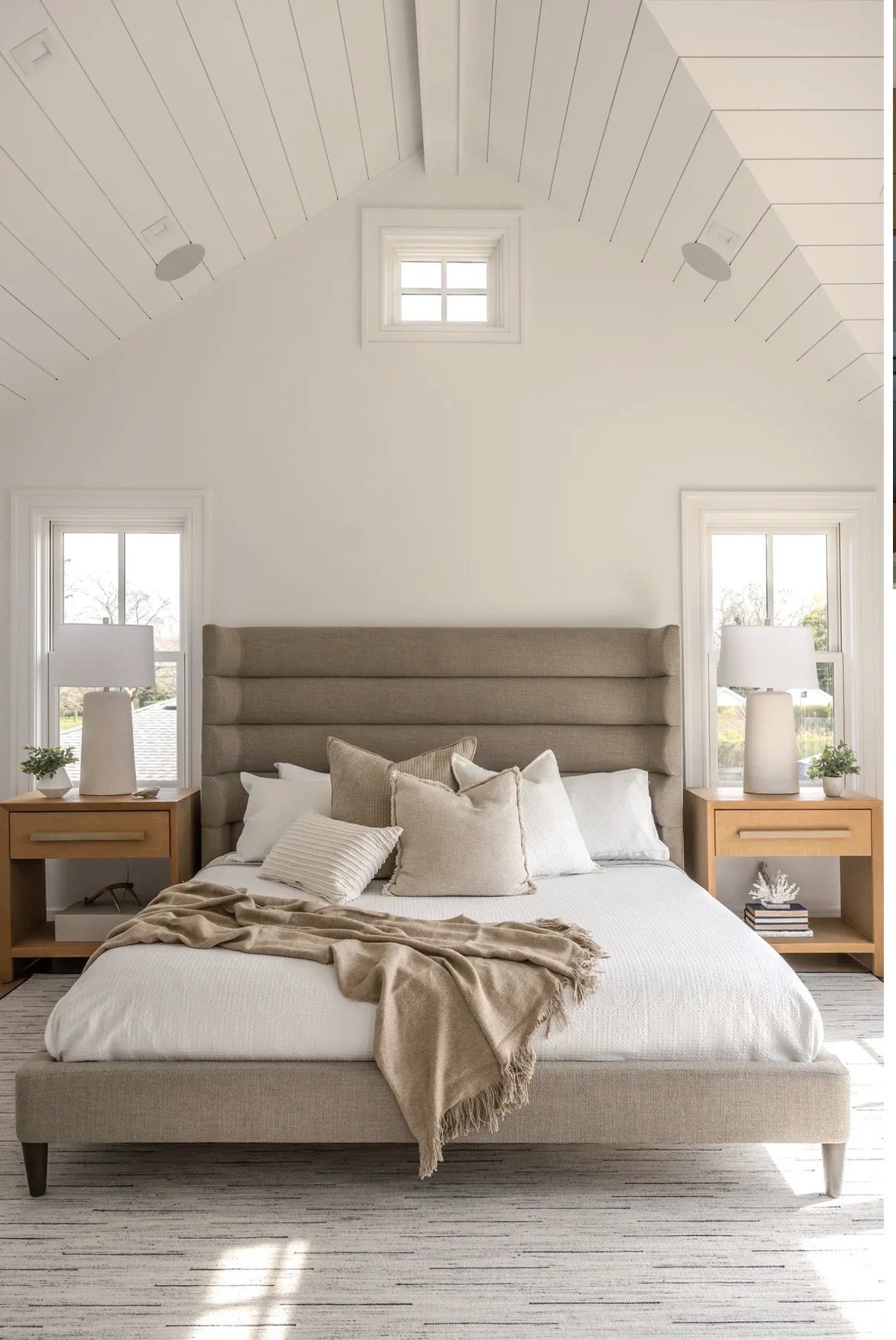 Modern bedroom design in Atlanta with a beige upholstered bed, layered pillows, and a textured throw. Featuring natural light, wood panel ceiling detail, and curated decor, this space blends warmth, comfort, and luxury interior styling.