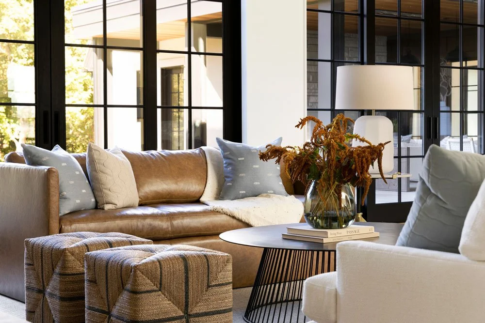 Modern Atlanta living room design with brown leather sofa, white accent chairs, and styled coffee table, featuring large black-framed windows, natural light, and indoor-outdoor living with patio views.