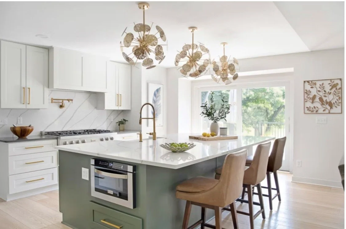 Luxury kitchen design in Atlanta featuring white cabinetry, a statement island with a green base, gold hardware, and glass pendant lighting. Large windows bring in natural light, creating a bright, modern space with upscale interior styling.