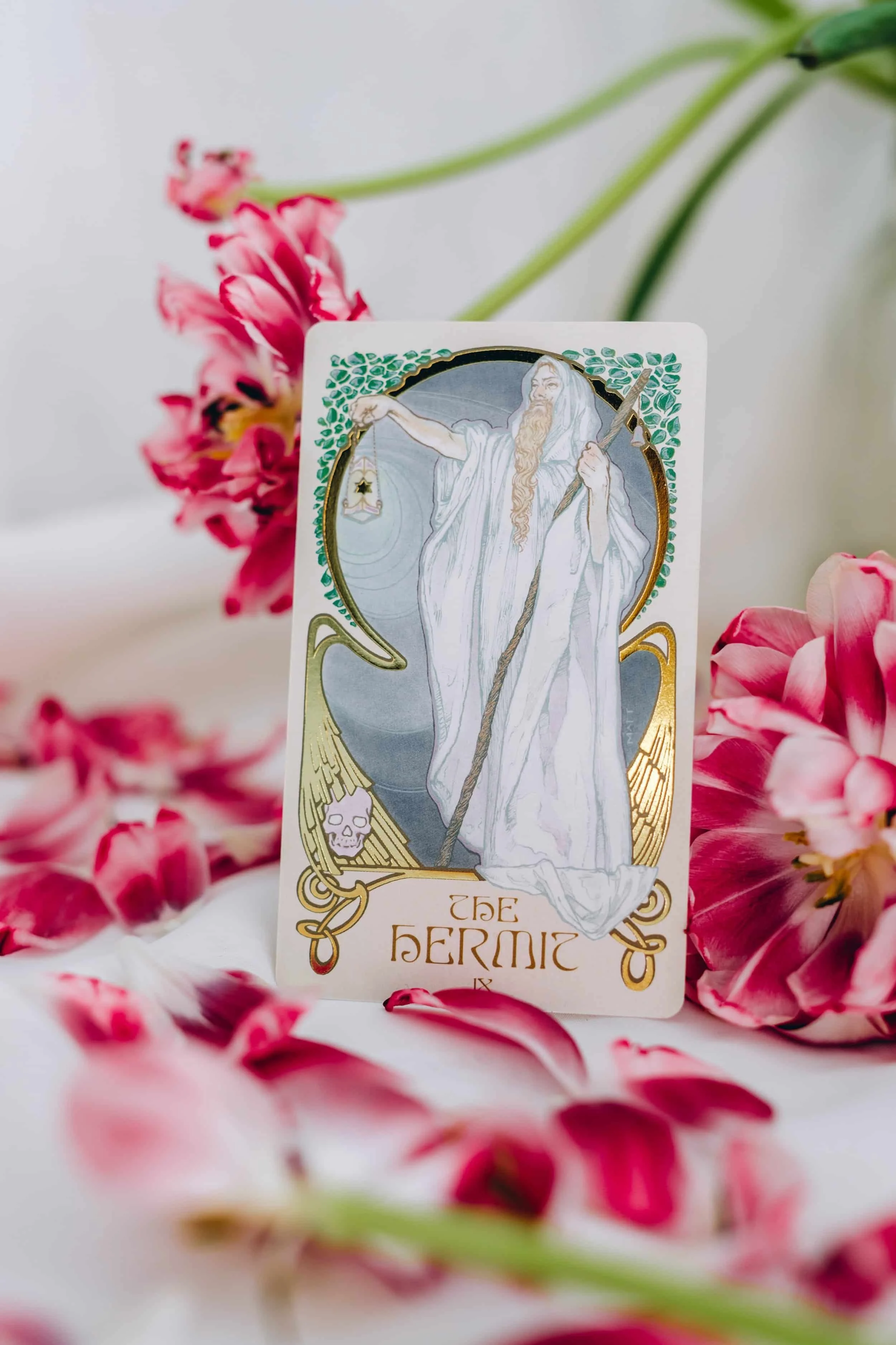 En tarotkort med bilden av en man i vita kläder, med ett långt skägg, som håller en stav och pekar med sin andra hand. Kortet har dekorativa element med gröna blad och gyllene detaljer, inklusive en liten skalle i hörnet.