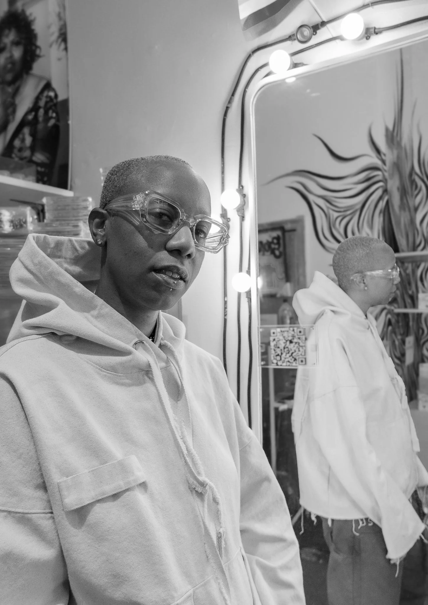 A woman with short hair and glasses standing in front of a mirror with lights, wearing a hoodie, with graffiti art in the background.