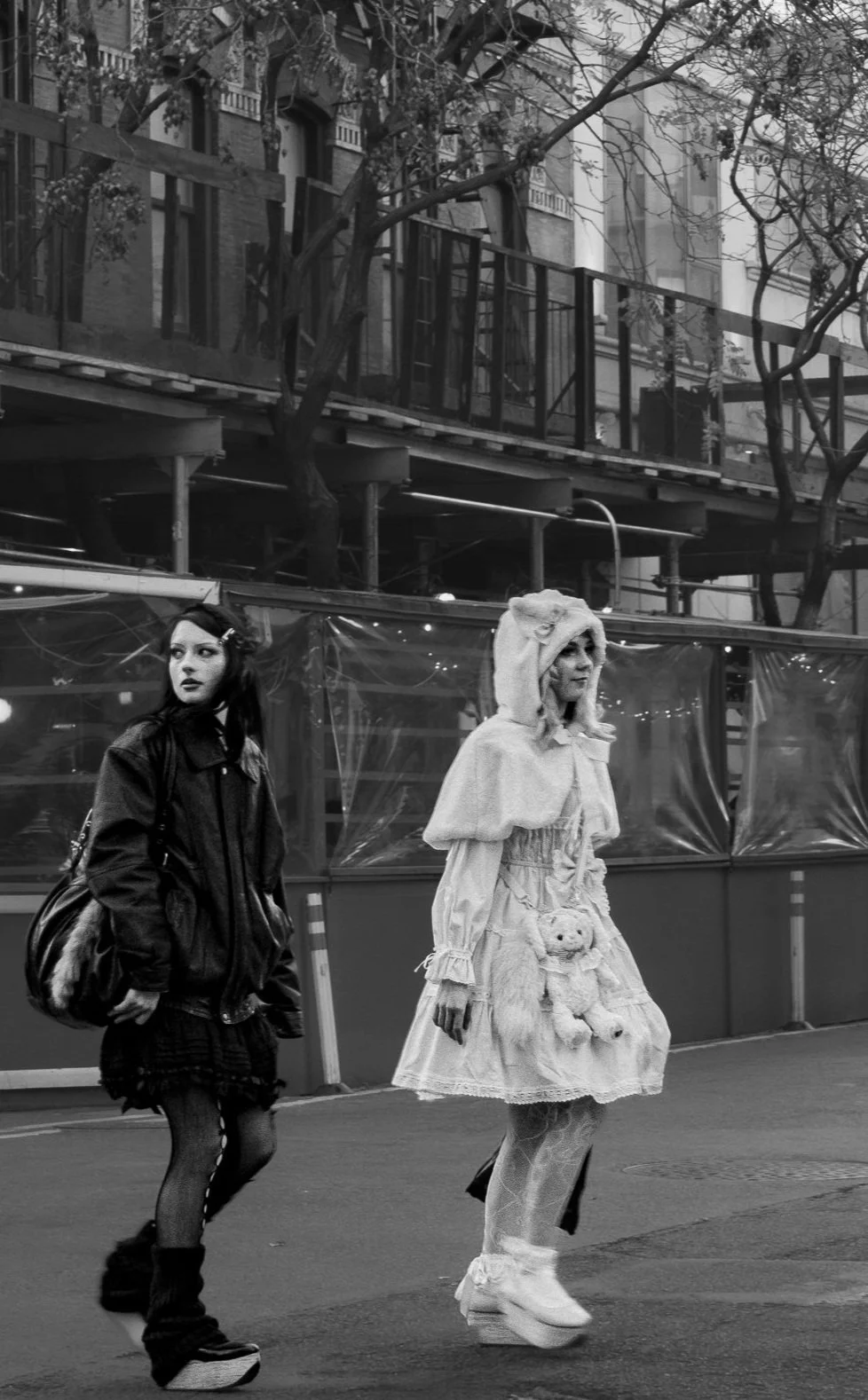 Two women walking on a city street; one in a dark jacket and the other in a light, whimsical dress with a hood and teddy bear accessories.