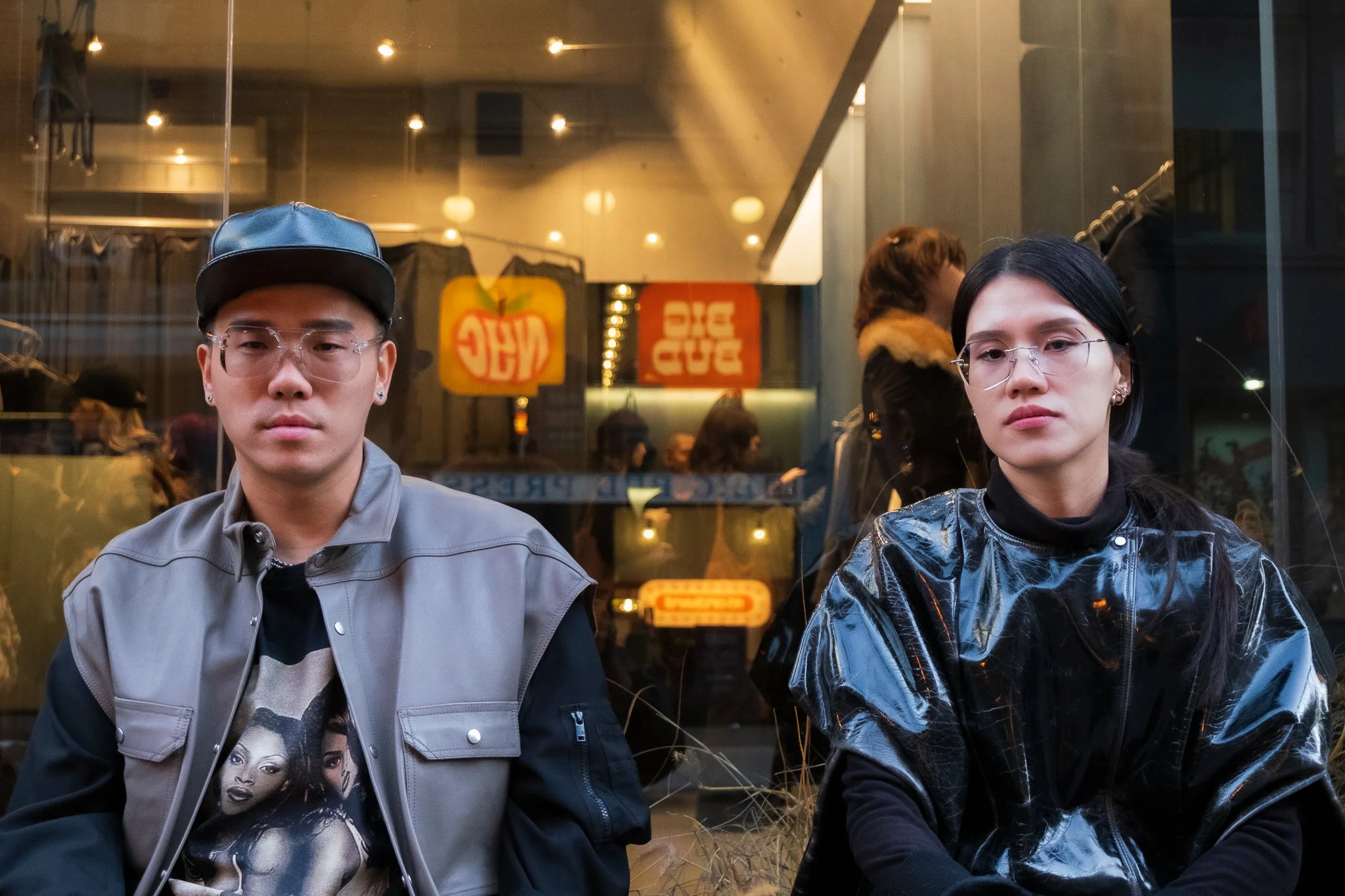 Two young adults sitting in front of a glass window at a restaurant or cafe during evening. The person on the left is wearing a gray jacket, a black cap, glasses, and a graphic t-shirt. The person on the right has long dark hair, glasses, and is wear