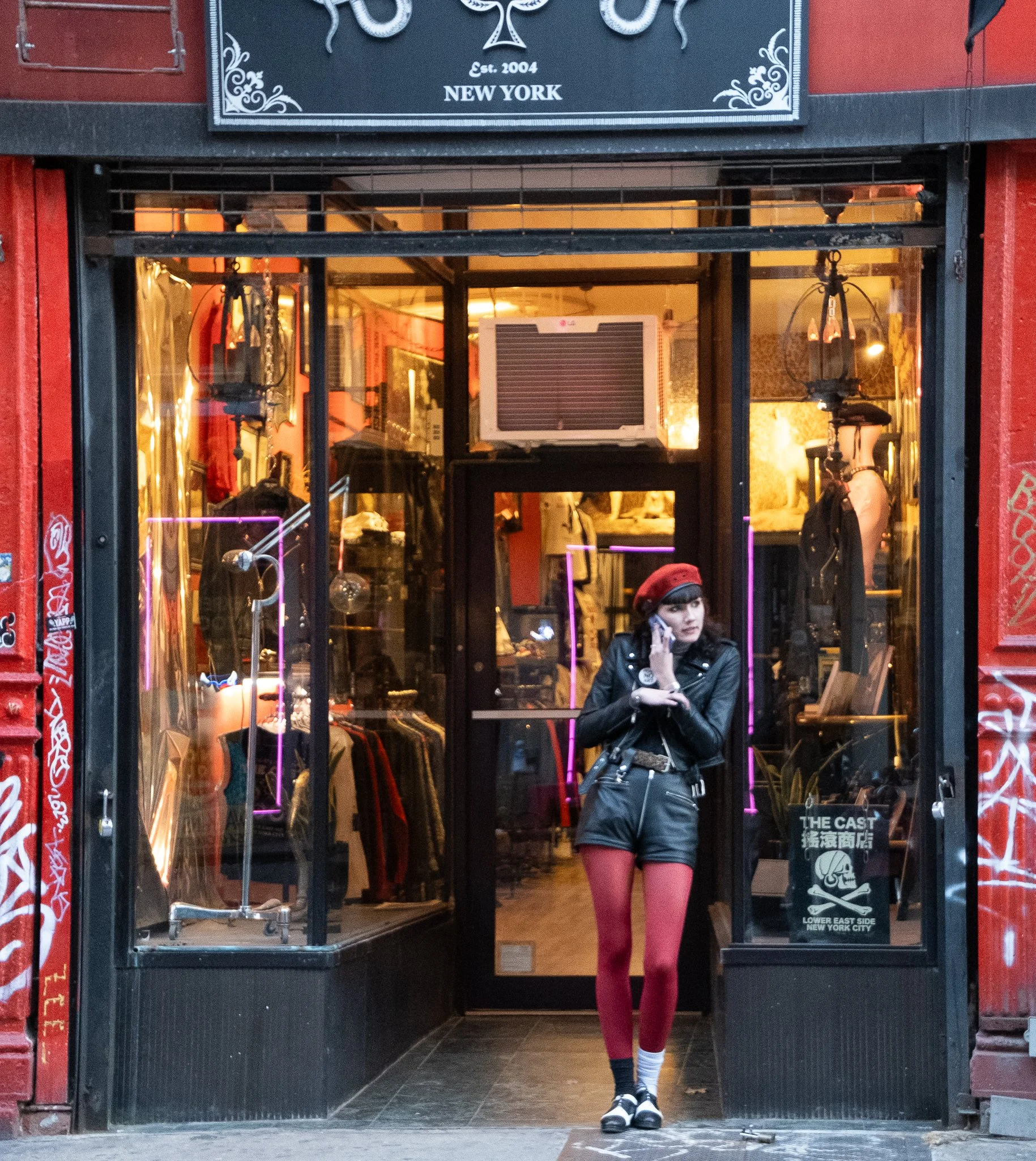 A young woman wearing red tights, a black leather jacket, and a red beret stands outside a clothing store on a city street, talking on her cellphone.